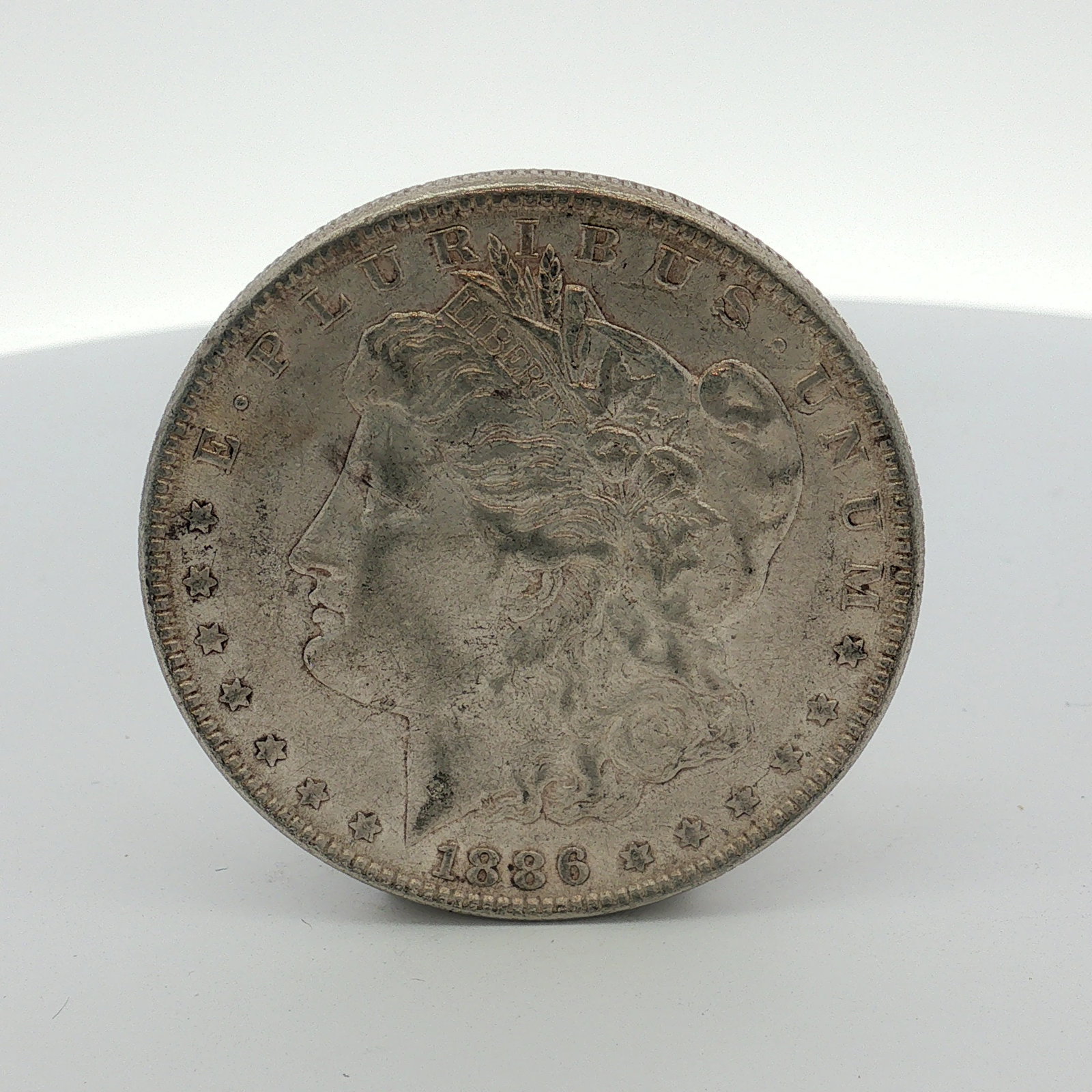 1886 US One Dollar Morgan Silver Dollar, Circulated: This 1886 US Morgan silver dollar was struck at the Philadelphia Mint. The coin is composed of 90% silver and 10% copper. It features the standard Morgan dollar design on the obverse and reverse. The 