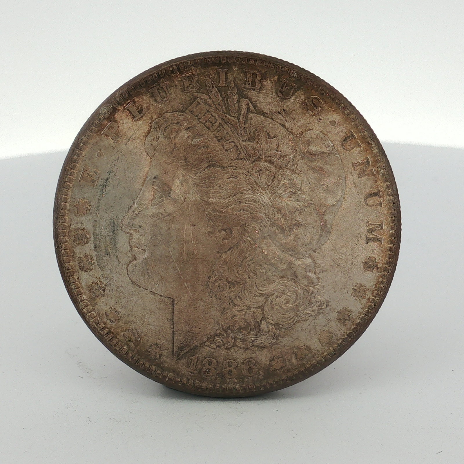 1886 US One Dollar Morgan Silver Dollar, Circulated: This US 1886 Morgan dollar is a struck silver coin produced at the Philadelphia Mint. It features the Liberty head obverse and the eagle reverse. The reverse displays an no mint mark below the wreath 