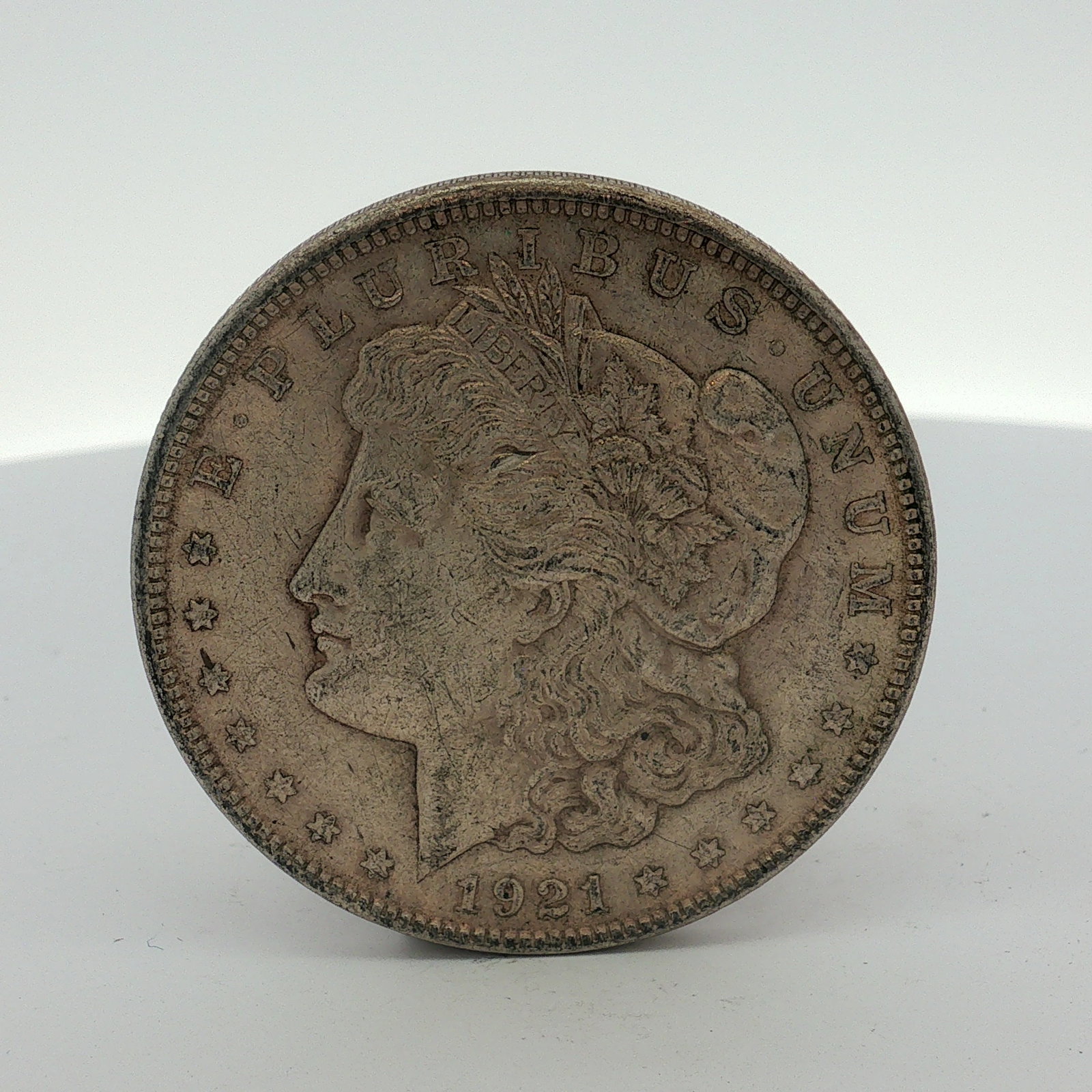 1921 US Morgan Silver Dollar Coin Circulated: This 1921 US Morgan Silver Dollar is a silver coin featuring the Morgan dollar obverse and reverse designs. The obverse is inscribed with E PLURIBUS UNUM, LIBERTY, and the date 1921. The reverse bears