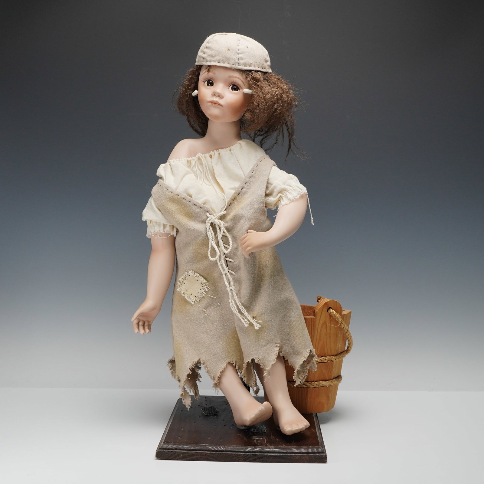 2pc Helen M. Carr Hamilton Collection Doll & Bucket, Ashley McTiddle: A finely crafted porcelain doll depicting a young girl in tattered period attire, posed & affixed on a wooden base with a rustic bucket accessory. The figure features expressive brown eyes,
