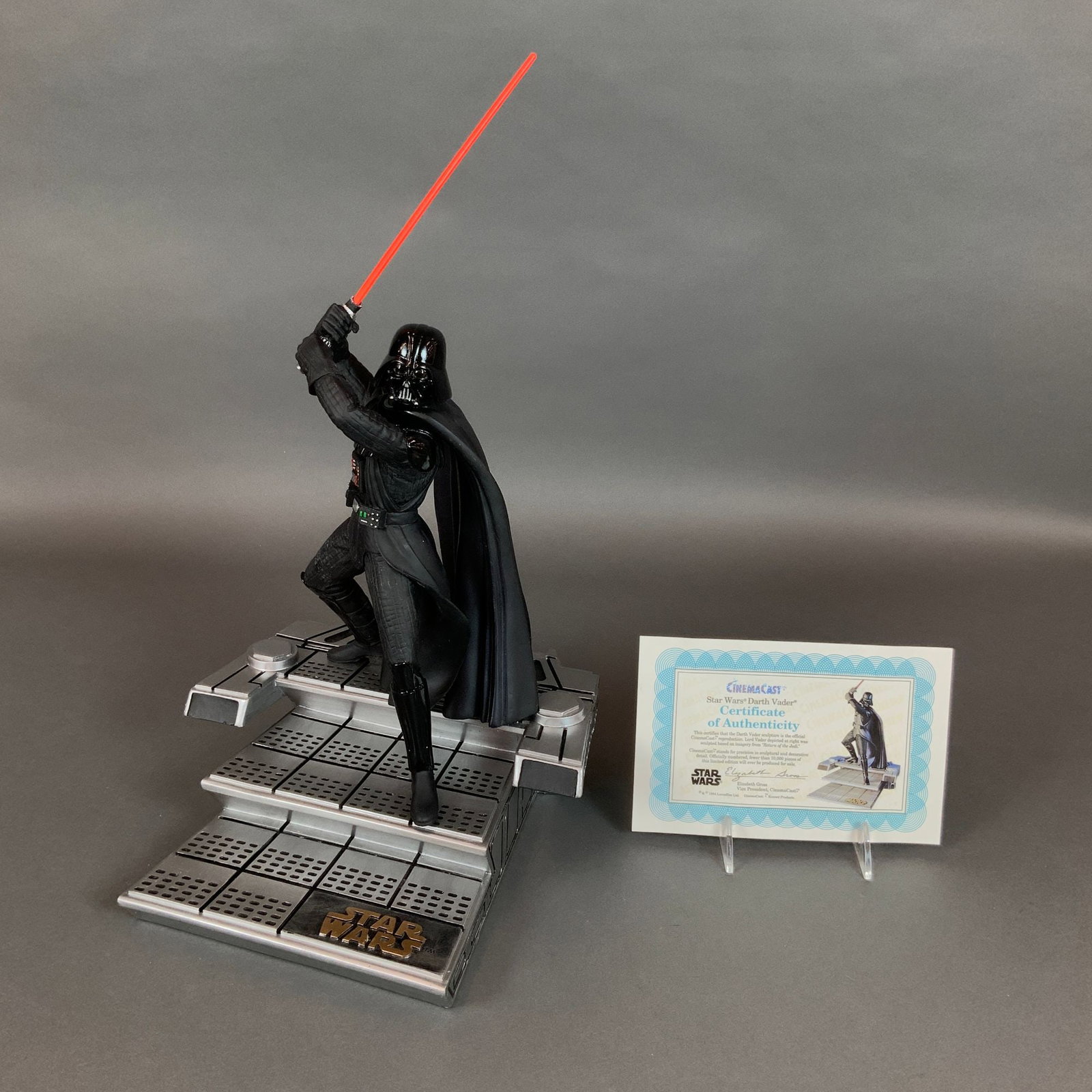 1994 CinemaCast Star Wars Darth Vader Limited Edition Sculpture: The piece depicts Darth Vader in a lunging pose with a raised red lightsaber, positioned on a metallic-finished base modeled after the interior of the Death Star throne room. The sculpture is construc