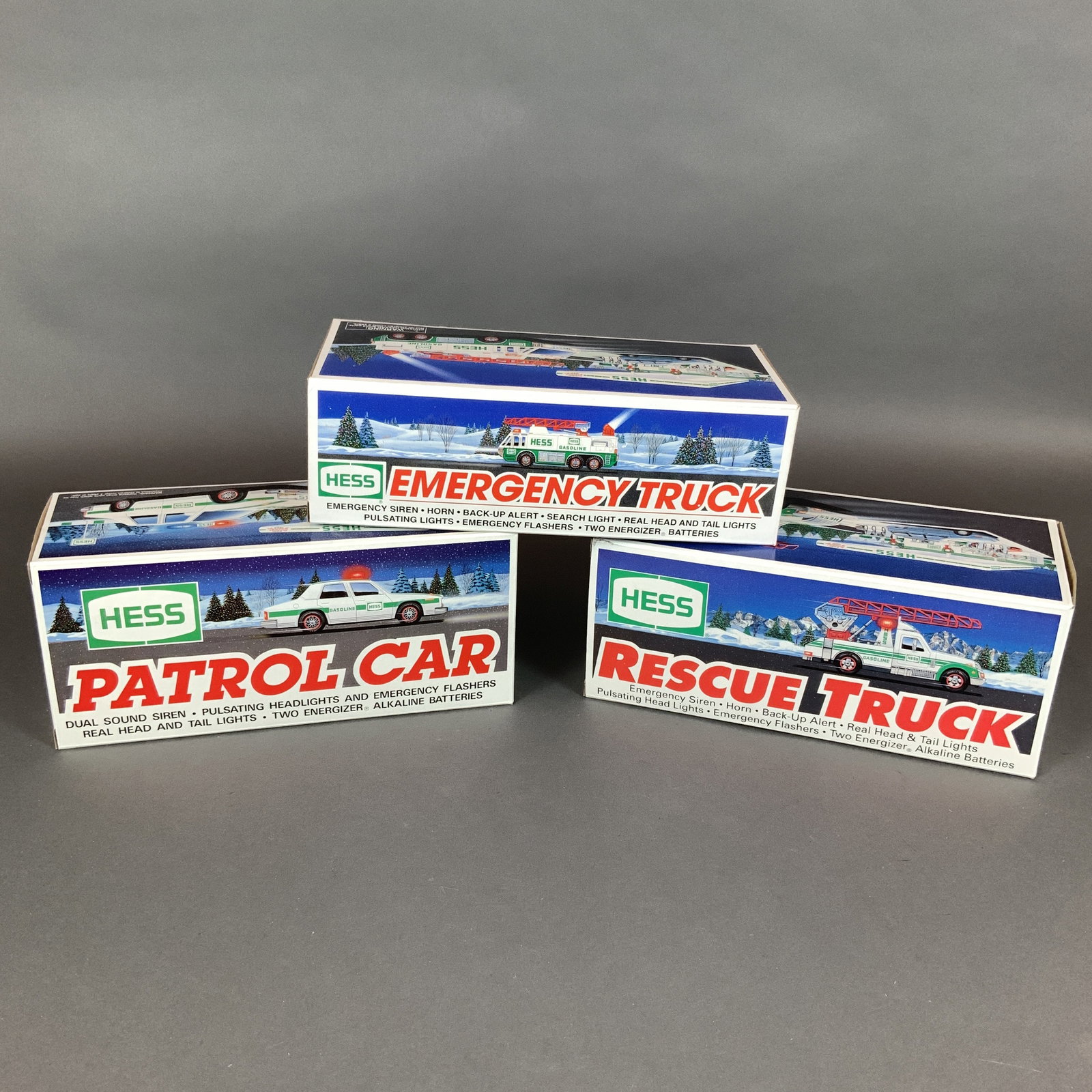 Three Hess Corporation Toy Vehicles in Original Boxes: A trio of vintage toy vehicles manufactured by the Hess Corporation, including the 1993 Patrol Car, the 1994 Rescue Truck, and the 1996 Emergency Truck. Each vehicle is constructed primarily of plasti