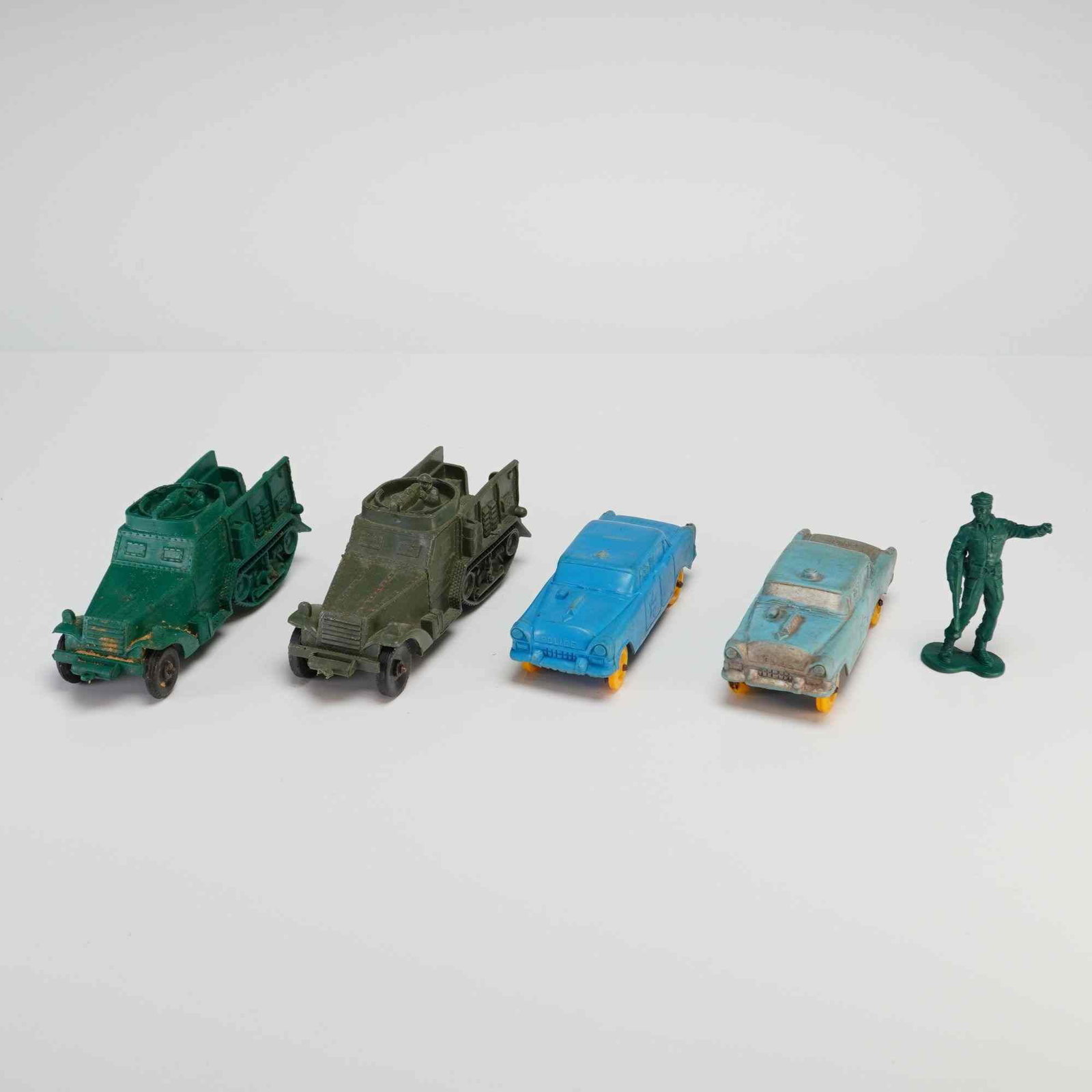 5pc Plastic Toy Army and Police Vehicles with Soldier Figure: This collection consists of vintage molded soft plastic toys, including military vehicles, police cars, and a soldier figure. The group features two military half-track vehicles, one in bright green a