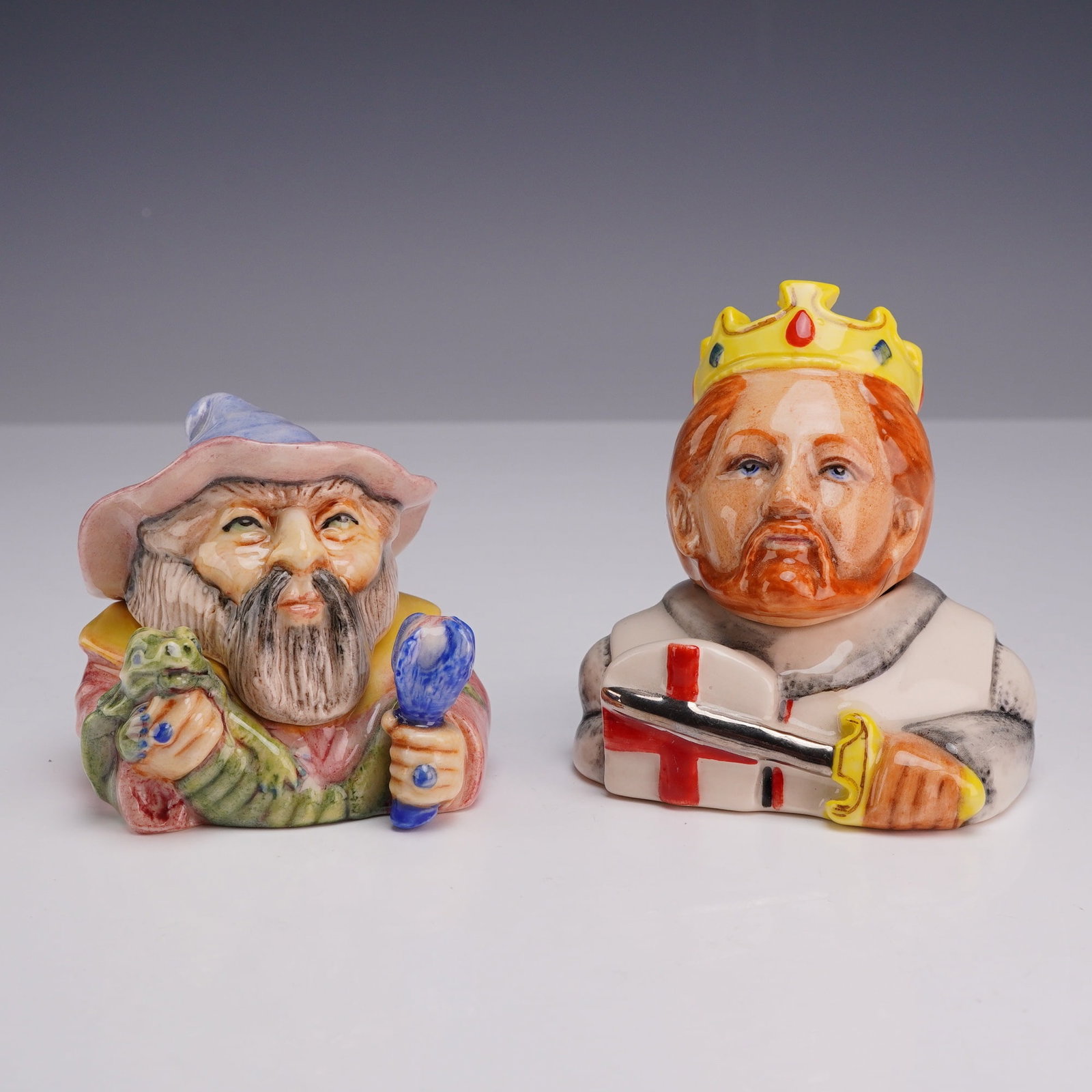 Two Kevin Francis Face Pots, Merlin and King Richard the Lionheart: These mold-formed pieces are hand-decorated with a high-gloss finish. The first piece represents Merlin the wizard, modeled with a gray beard and blue pointed hat, holding a green dragon and a blue st