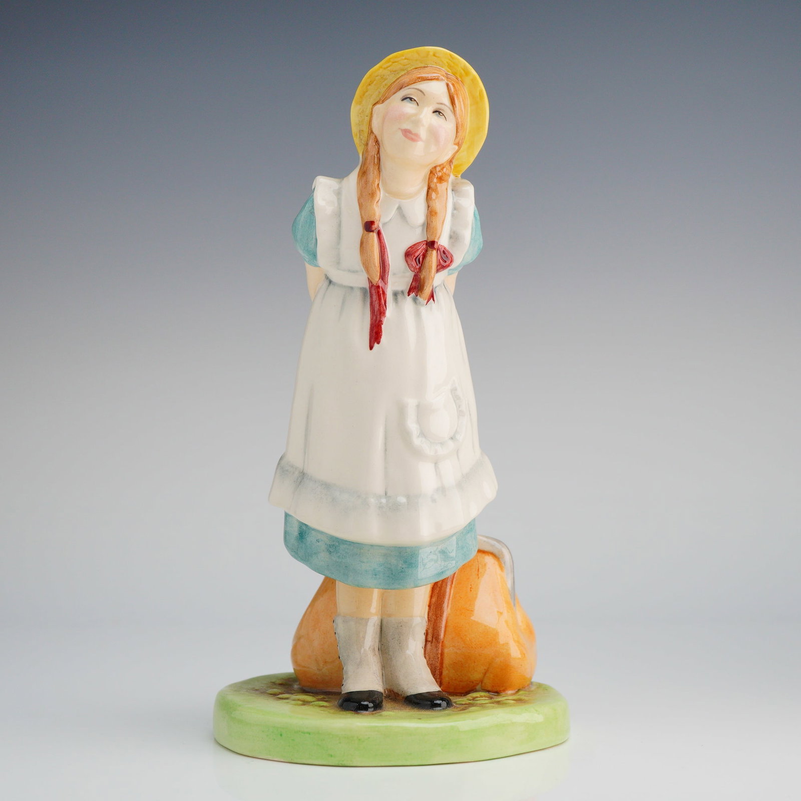 Royal Doulton Figurine, Pollyanna HN2965: The figure depicts a young girl standing on a circular grassy base, wearing a light blue dress, white apron, and a yellow straw hat, with her hands clasped behind her back. She stands next to an orang
