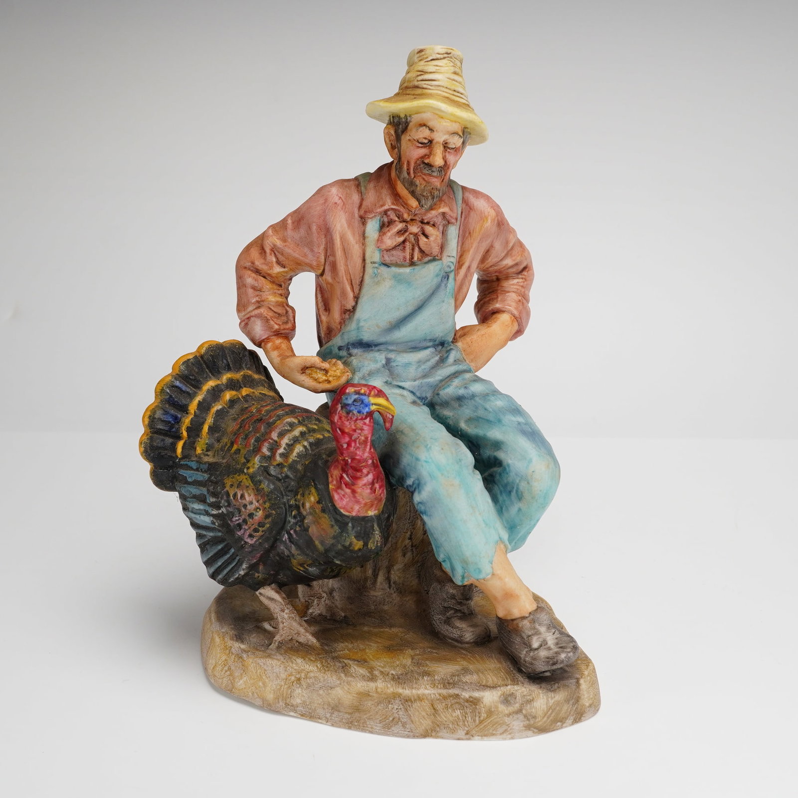 Royal Doulton Figurine, Thanksgiving HN2446: A Royal Doulton porcelain figurine titled Thanksgiving, model number HN2446, featuring a matte finish. The piece depicts a seated farmer wearing a yellow straw hat, a pink shirt, and blue overalls. Th