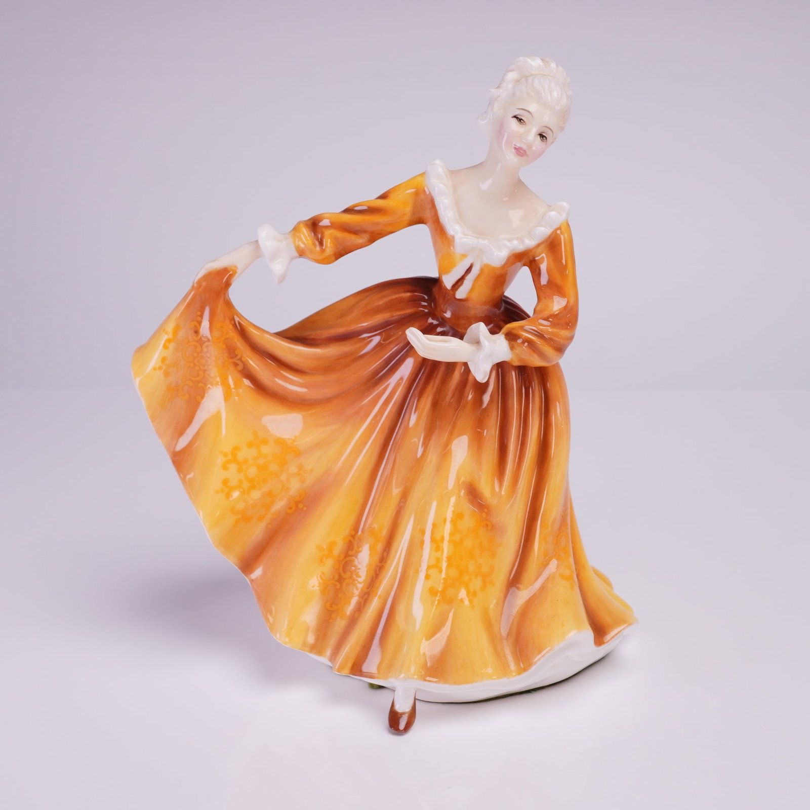 Royal Doulton Figurine Kirsty HN2381: The figure depicts a woman in a dancing or curtseying pose, lifting the voluminous skirt of her gown. The dress is hand-painted in high-gloss shades of amber and burnt orange, featuring subtle lighter