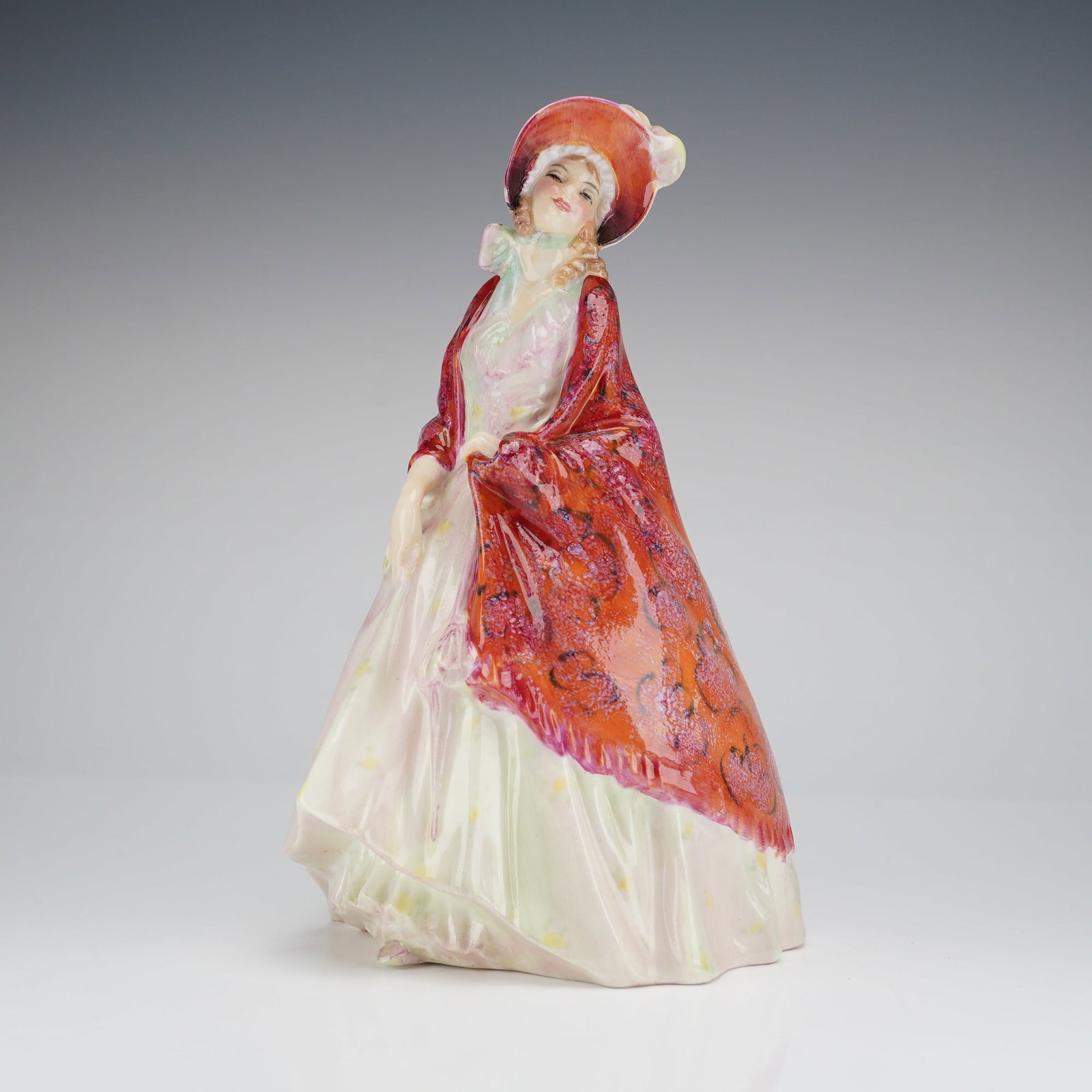 Royal Doulton Figurine Paisley Shawl HN 1392: The figure depicts a woman modeled in a period style, characterized by a long, flowing pale dress and a large, high-relief red shawl detailed with a dark paisley pattern. The piece is rendered in vibr