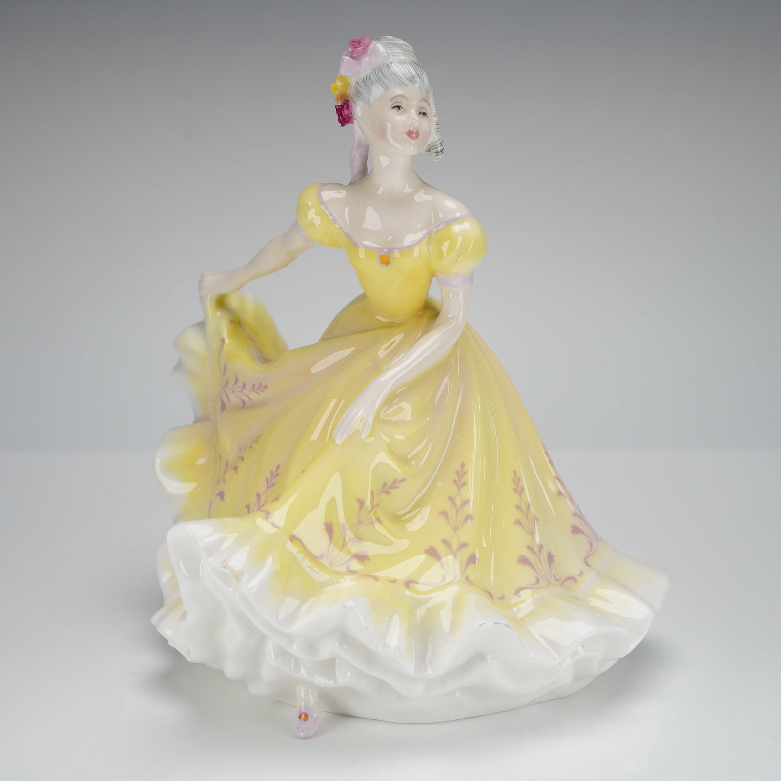 Royal Doulton Figurine, Ninette HN2379: The figure depicts a woman in a dynamic pose suggestive of dancing or curtsying, dressed in a period-style gown. The dress is predominantly glazed in vibrant yellow, detailed with hand-painted mauve f