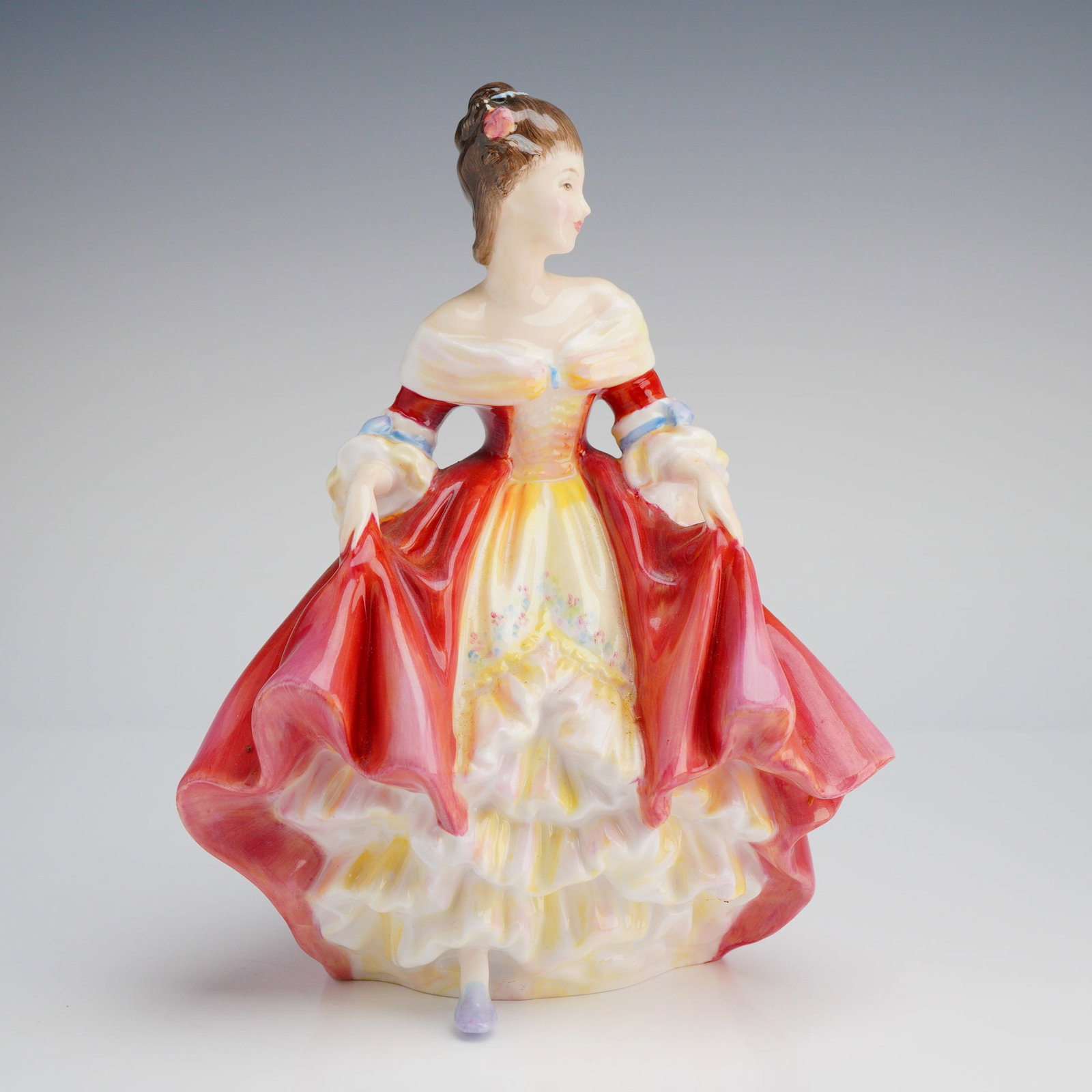 Royal Doulton Figurine, Southern Belle HN2229: The figure is hand-painted and glazed, depicting a woman in a dramatic period gown characterized by a voluminous red overskirt lifted by her hands to reveal a tiered yellow and white ruffled underskir
