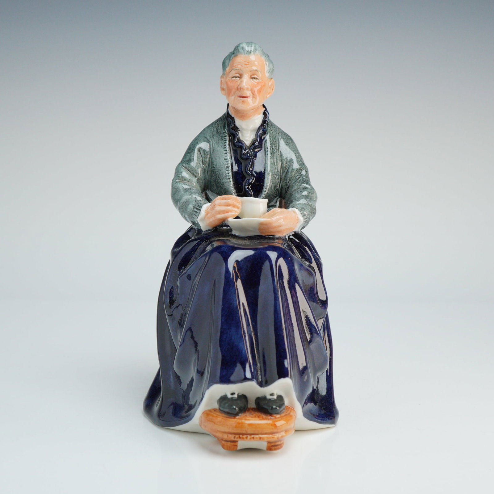 Royal Doulton Figurine, The Cup of Tea HN2322: Part of the Golden Year series, figure is sitting enjoying her drink, modeled by Mary Nicoll. Colored in dark blue and gray. Royal Doulton backstamp. Dimensions: 4.50"L x 3.25"W x 6.75"H Weight: 1.09 