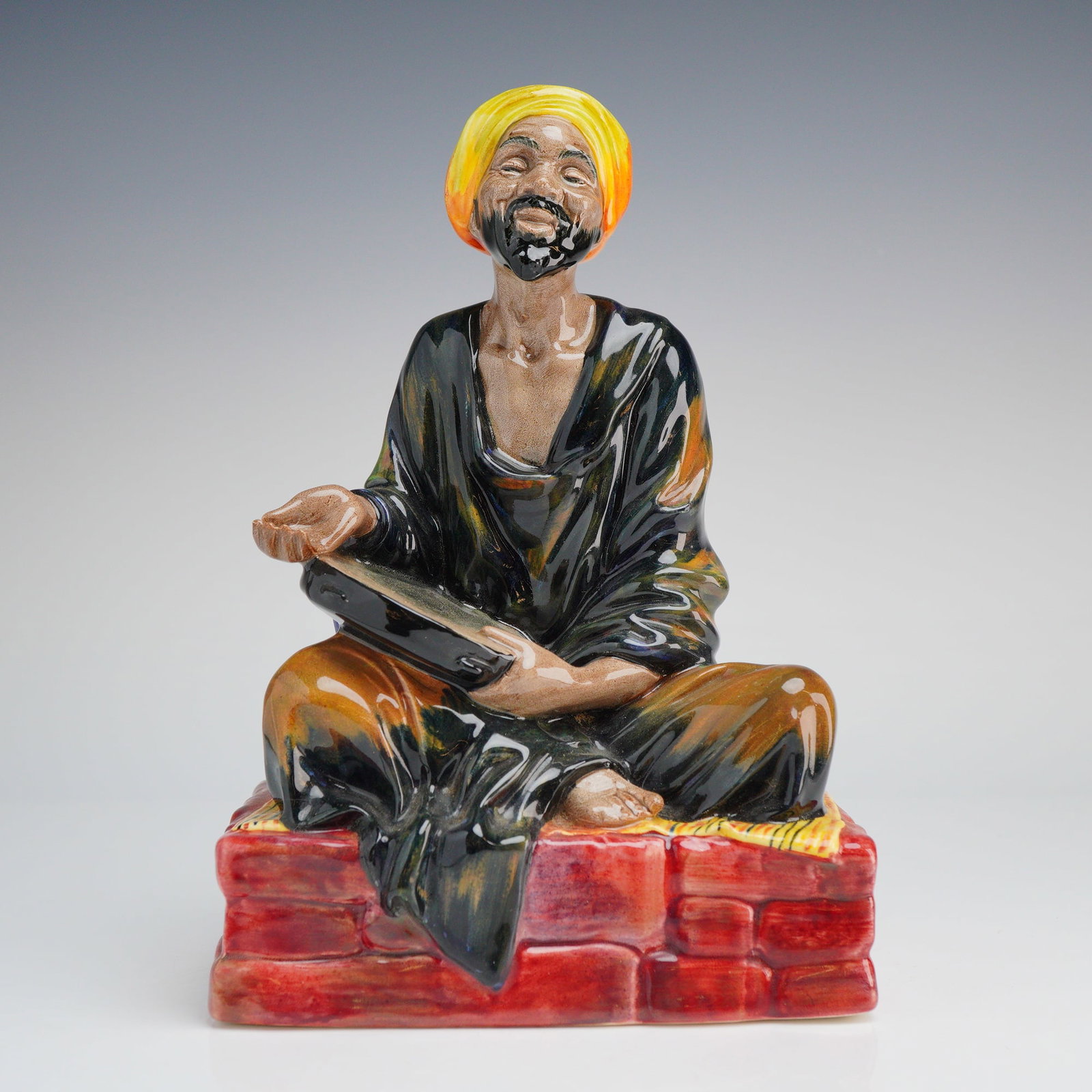 Royal Doulton Porcelain Figurine, Mendicant HN1365: A Royal Doulton glazed porcelain figurine titled The Mendicant, identification number HN 1365. The figure depicts a man with a black beard and a yellow and orange turban, seated cross-legged on a red 