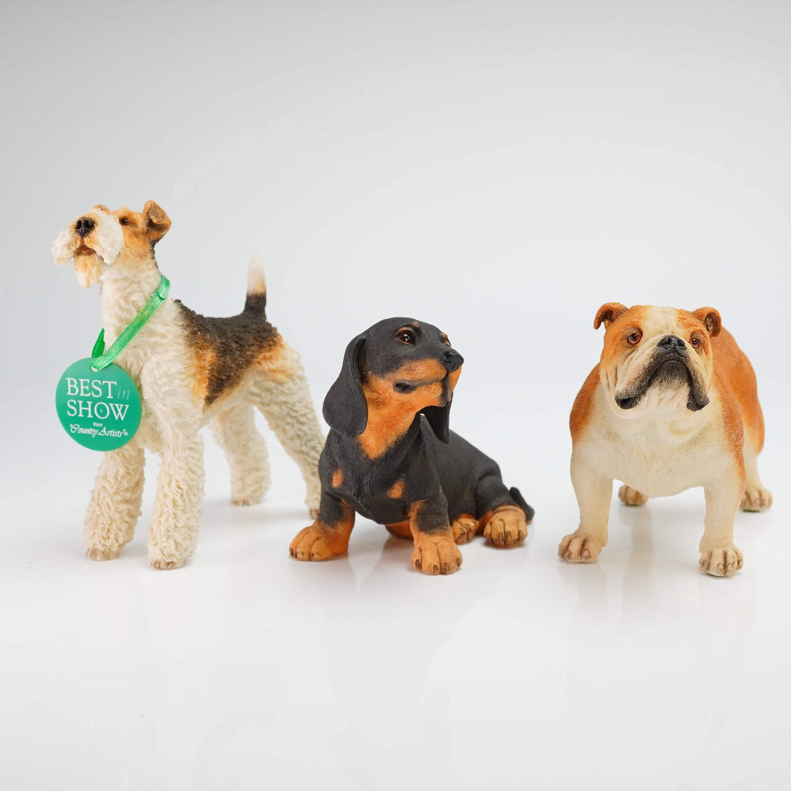 Set of Three Country Artists England Resin Dog Figurines, Best in Show: A collection of three hand-painted cast resin dog figurines produced by Country Artists. The group includes a wire-haired fox terrier, a black and tan dachshund puppy, and an English bulldog. Each fig