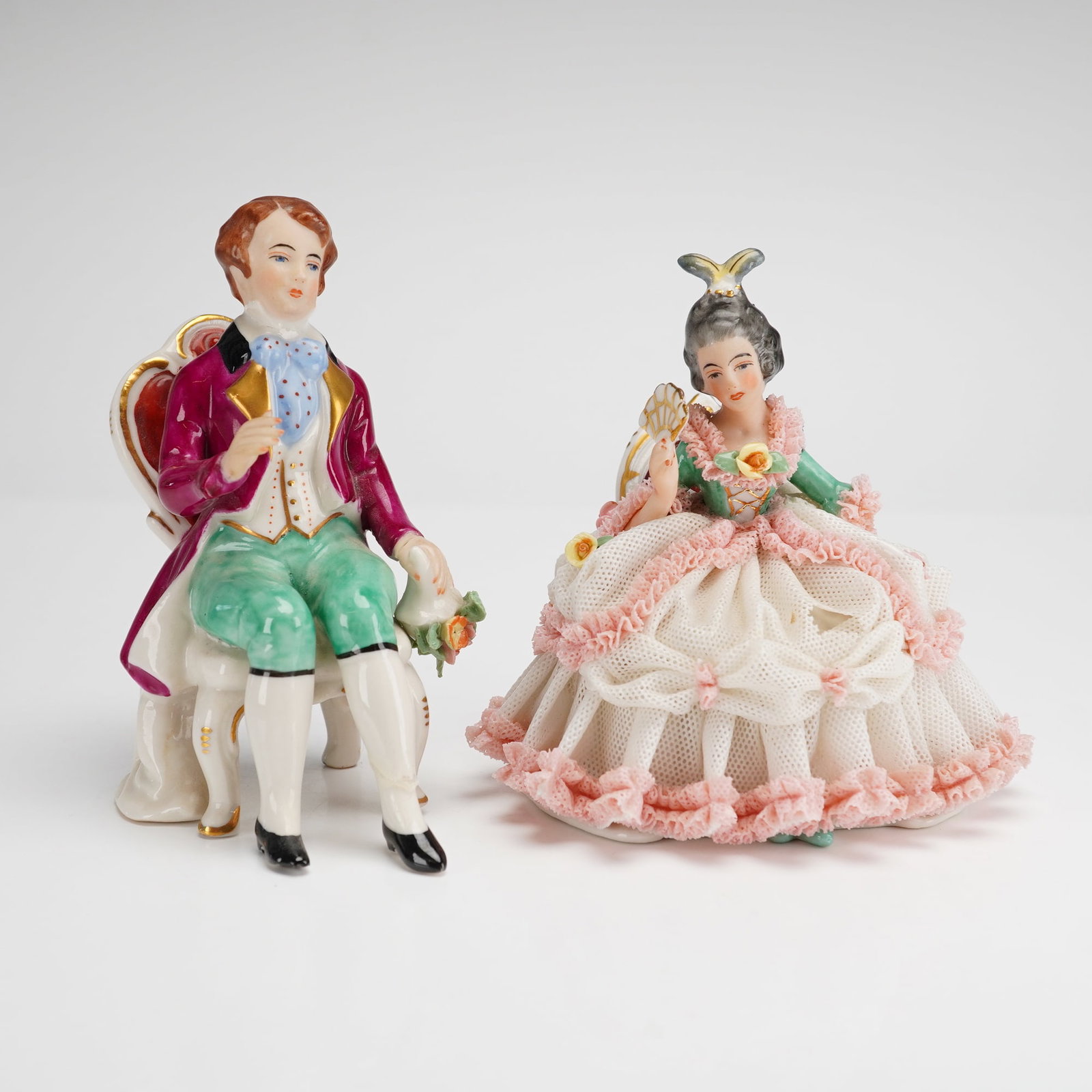 2pc Frankenthal Wessel Porcelain Lace Figurines: Porcelain lace figurines are hand-painted and feature intricate applied porcelain lace details. The male figure is depicted in a purple frock coat and green breeches, seated on a gilt-accented chair w