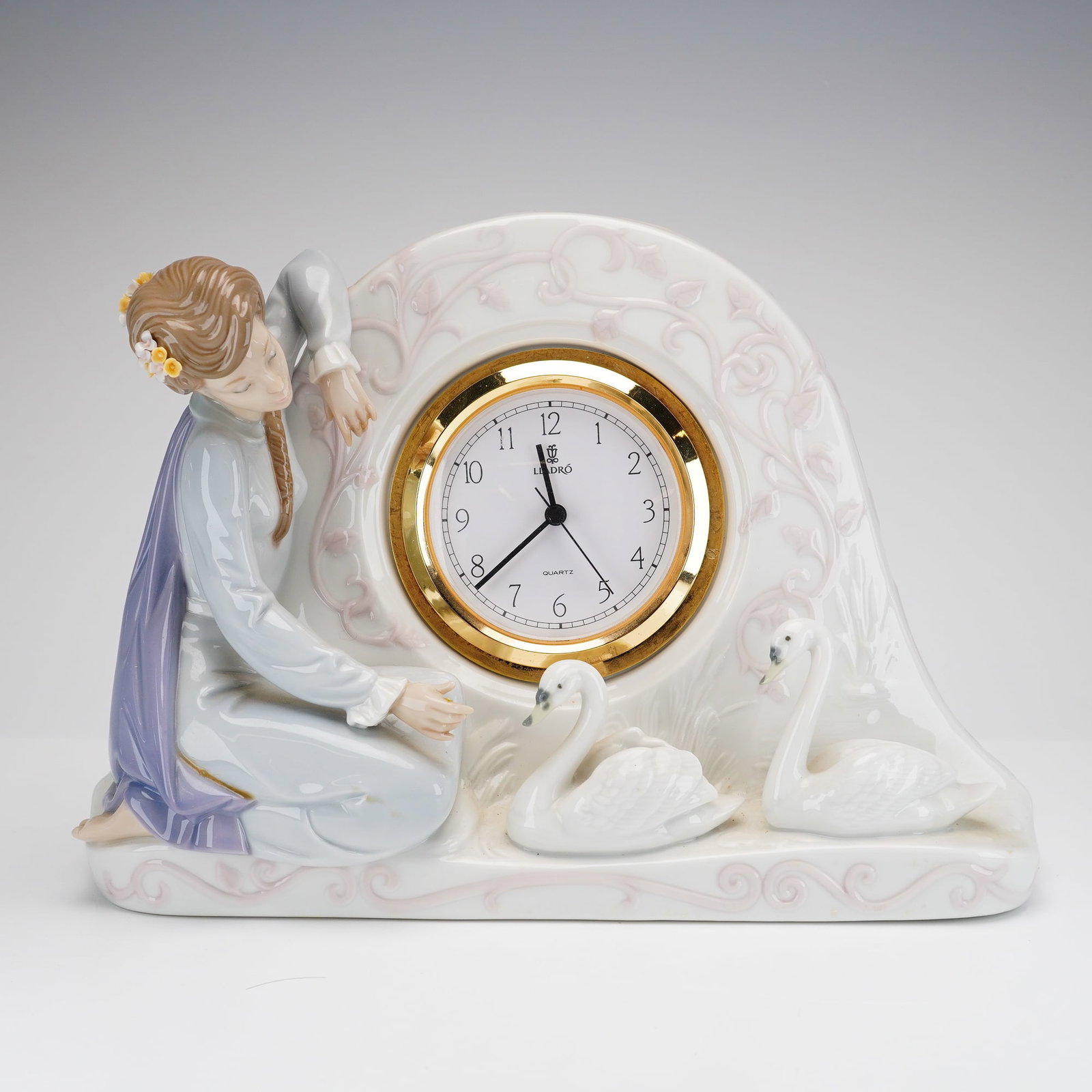 Lladro Porcelain Swan Clock 01005777: This polychrome glazed porcelain piece features an arched body adorned with raised, bas-relief scrolling vines and leaf motifs in a pale lavender-pink glaze against a white ground. To the left of the 