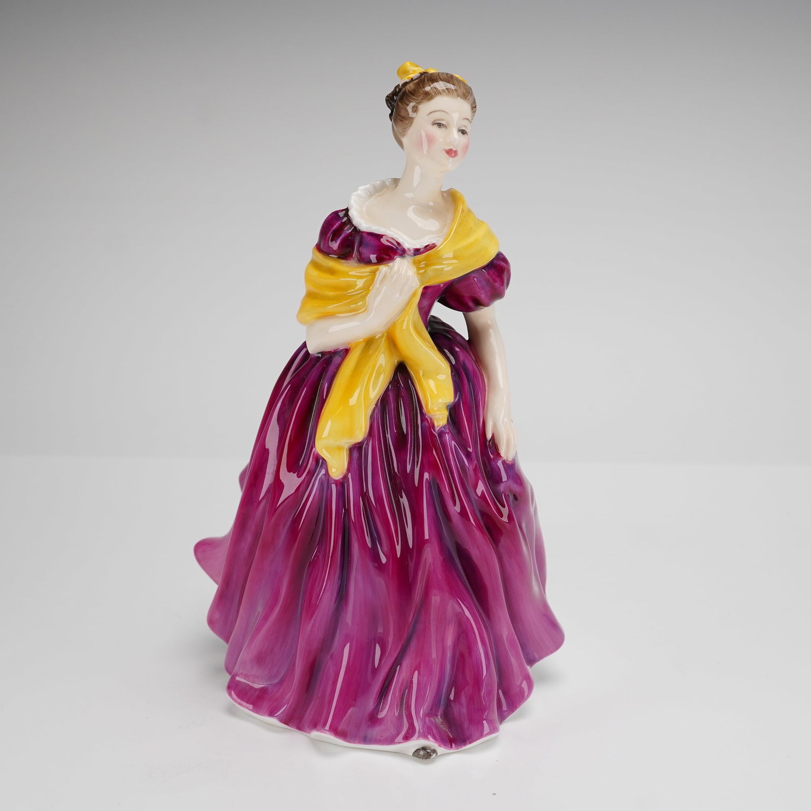 Royal Doulton Adrienne Figurine HN2152: A Royal Doulton bone china figurine titled Adrienne, model number HN2152. The figure is depicted in a standing pose wearing a voluminous magenta gown and a draped yellow shawl. The hair is gathered an
