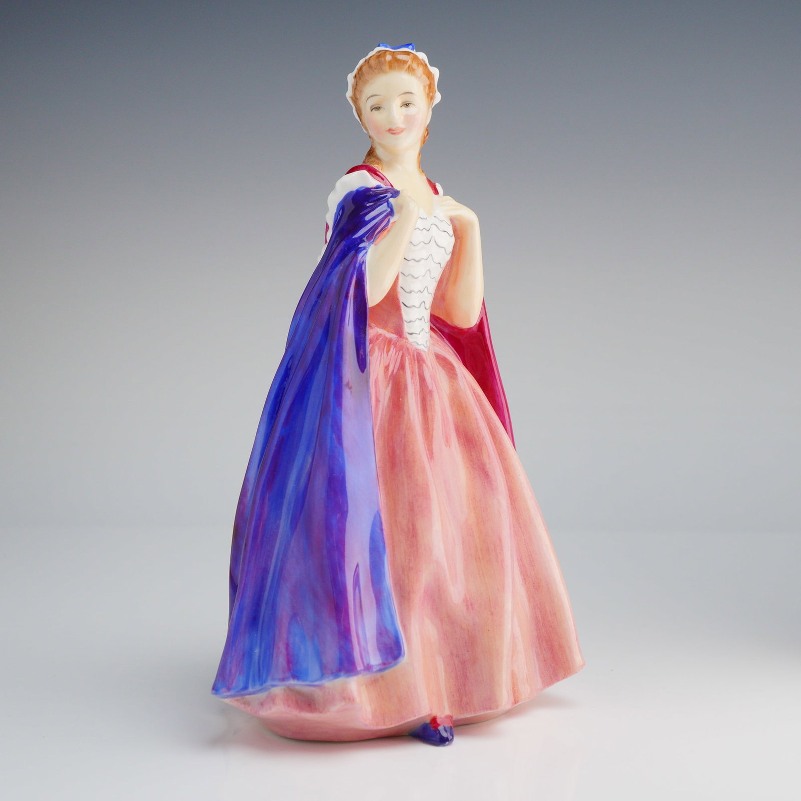 Royal Doulton Figurine, Bess HN2003: The hand-painted ceramic figure depicts a young woman in a full-length, peach-pink gown, a white bodice, and a white bonnet accented with blue. She is shown adjusting a dramatically draped cloak, whic
