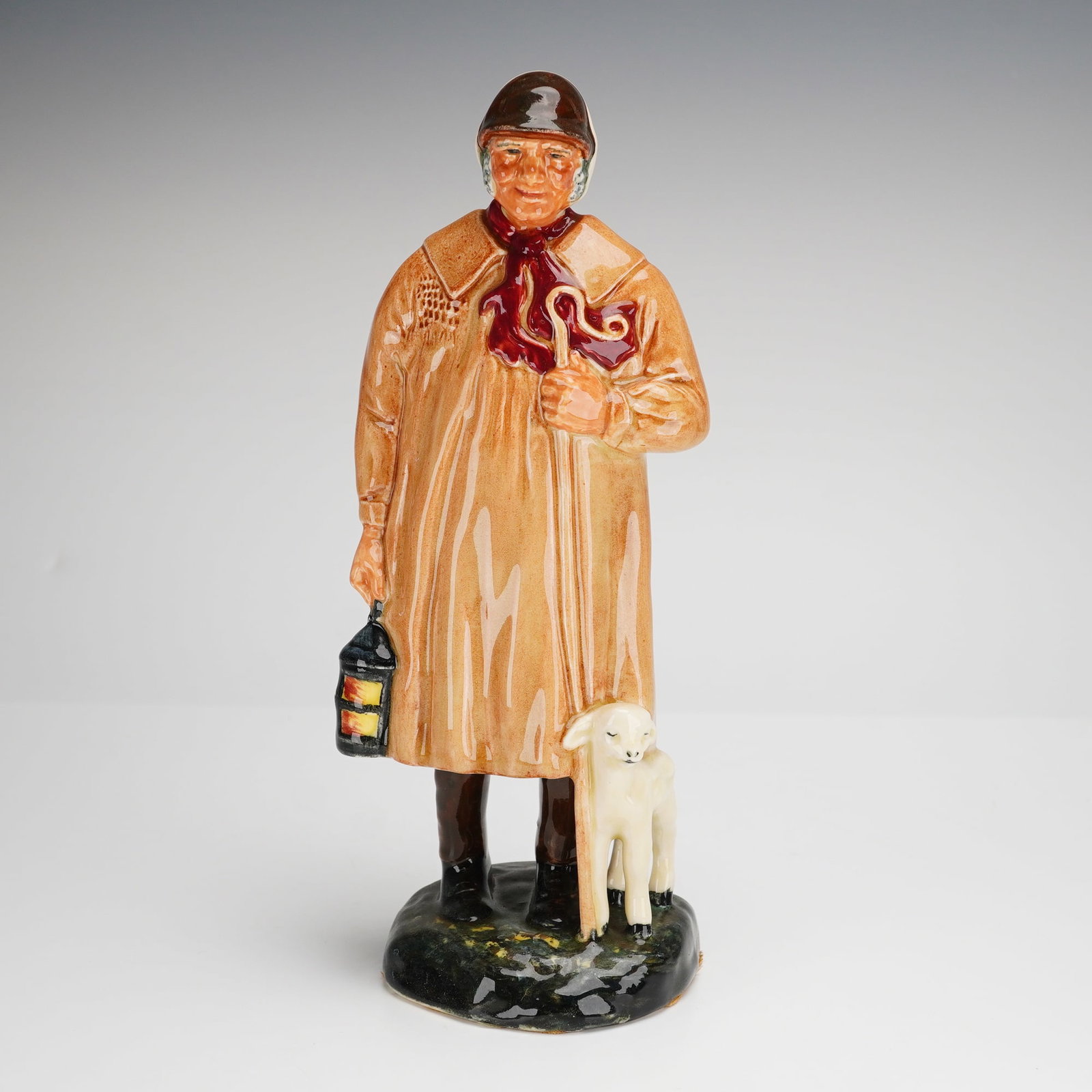 Royal Doulton The Shepherd HN1975 Figurine: A Royal Doulton glazed ceramic figurine titled The Shepherd, model number HN1975. The piece depicts a standing male figure in a long tan smock with a red neckerchief and brown cap, holding a shepherd'