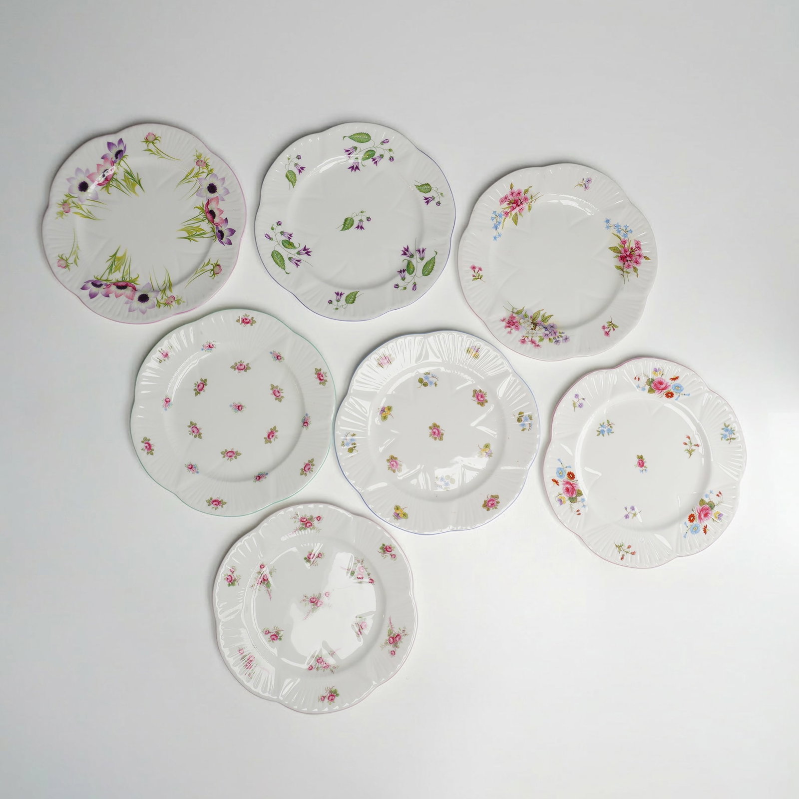 7pc Shelley Fine Bone China Dainty Plates: This set of seven fine bone china plates is manufactured by Shelley in England. The plates feature the Dainty shape, which is defined by a circular form with a scalloped and radially fluted rim. The d