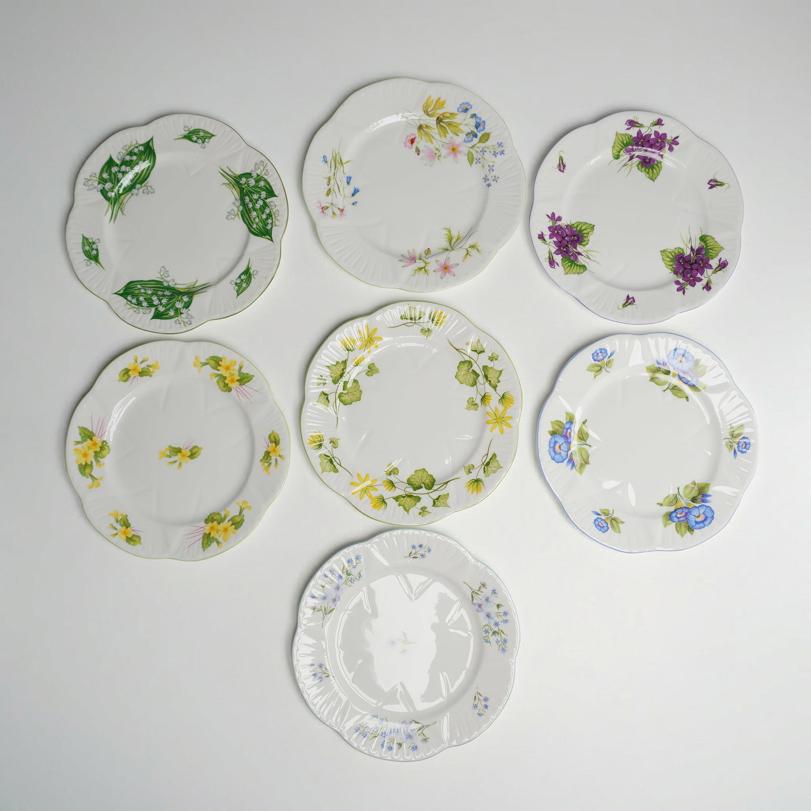 7pc Shelley Fine Bone China Dainty Shape Floral Plates: This collection of fine bone china plates was manufactured by Shelley in England. Each plate is designed in the Dainty shape, characterized by a deeply scalloped and fluted rim. The set features vario