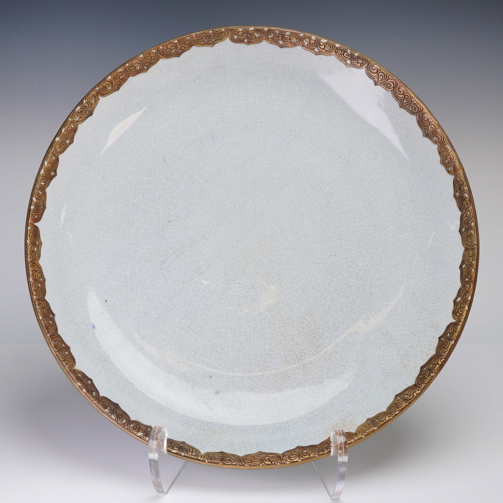 Metal-Mounted Crackle-Glazed Ceramic Charger: A circular charger covered in a pale light white glaze exhibiting extensive, fine crackling across the entire surface. The plate is distinguished by a heavy, applied metal mount, which forms the rim t