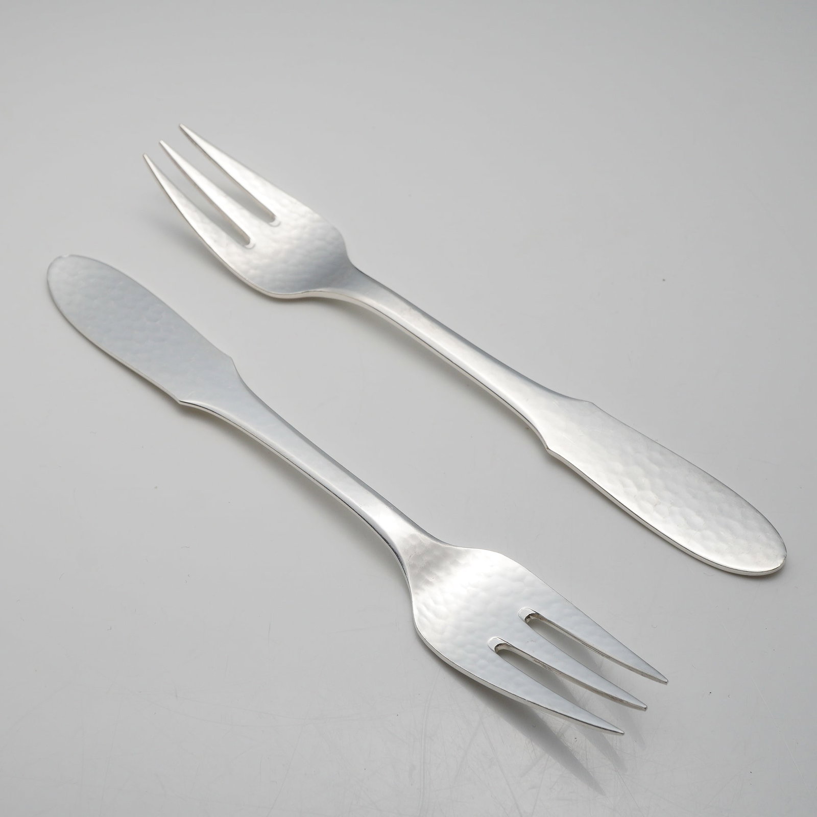 A Pair of Georg Jensen Denmark EPNS Mermaid Pattern Hammered Fish Forks: This pair of fish forks is manufactured by Georg Jensen of Denmark in the Mermaid pattern. The items are constructed from electroplated nickel silver, as indicated by the EPNS designation. The design 