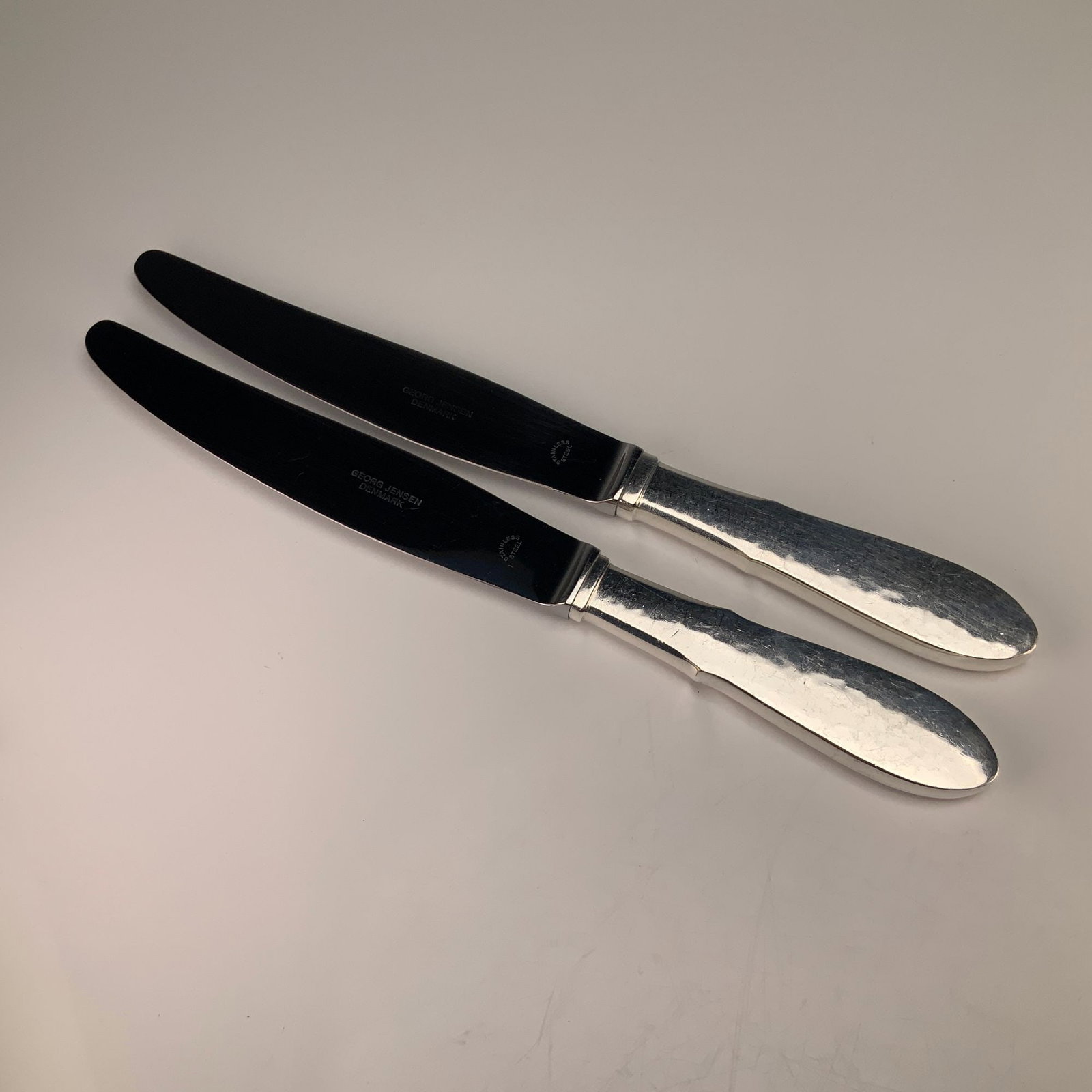 A Pair of Georg Jensen Denmark Mermaid Pattern EPNS Knives: This pair of knives is manufactured by Georg Jensen of Denmark in the Mermaid pattern. The pieces feature silver-plated handles with a hand-hammered finish and flared terminals. The handles are constr