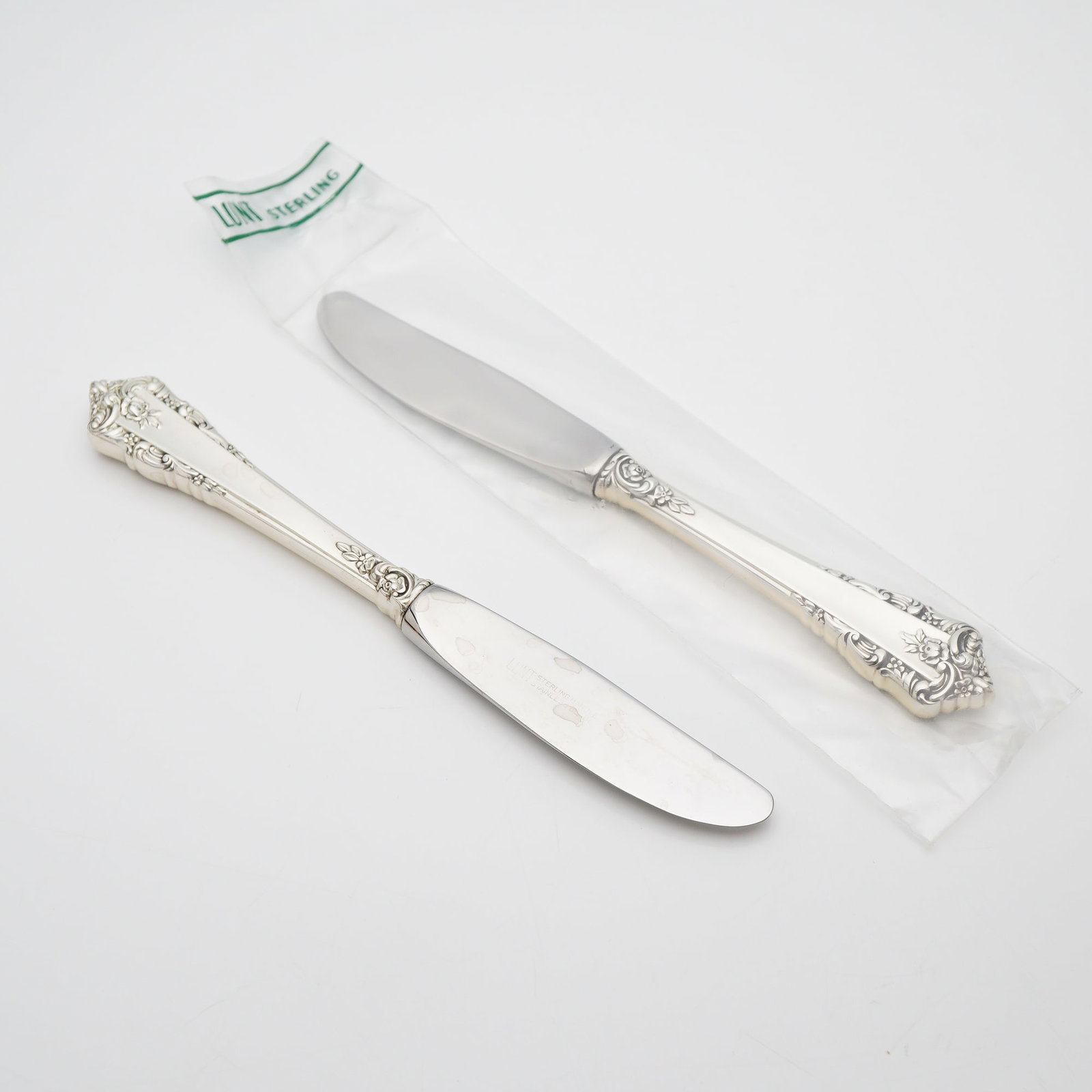2pc Lunt Sterling Silver and Stainless Steel Butter Spreaders Knives, Rondelay: These pieces feature sterling silver handles decorated with intricate scrolled edges and floral motifs. The spreaders are fitted with stainless steel blades. The blades are marked LUNT STERLING HANDLE