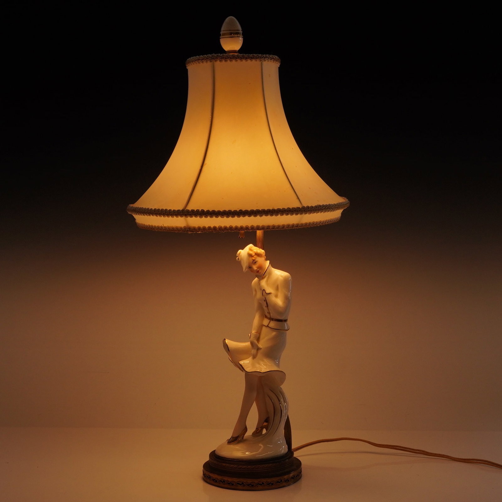 Vintage Art Deco Lamp with Porcelain Lady Figurine: This vintage Art Deco lamp with porcelain lady figurine features a glazed porcelain central figure of a woman in a stylized pose. The figurine is detailed with gilt accents on the shoes, belt, and dre