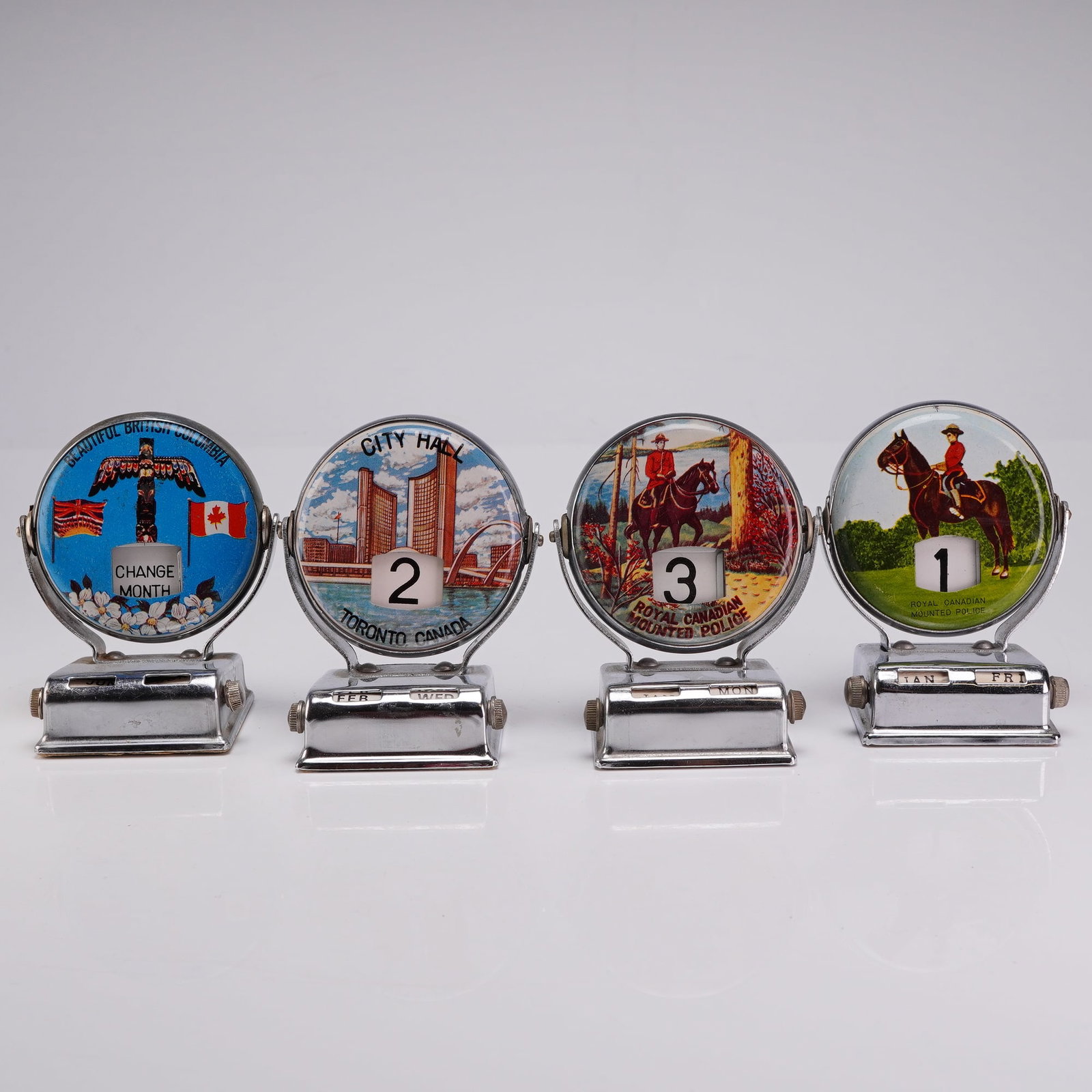 Set of Four Vintage Canadian Souvenir Roll Top Perpetual Flip Desk Calendars: These souvenir mechanical desk accessories are constructed from chrome-plated metal with clear plastic lenses and felt-lined bases. The date advances automatically when the circular housing is rotated