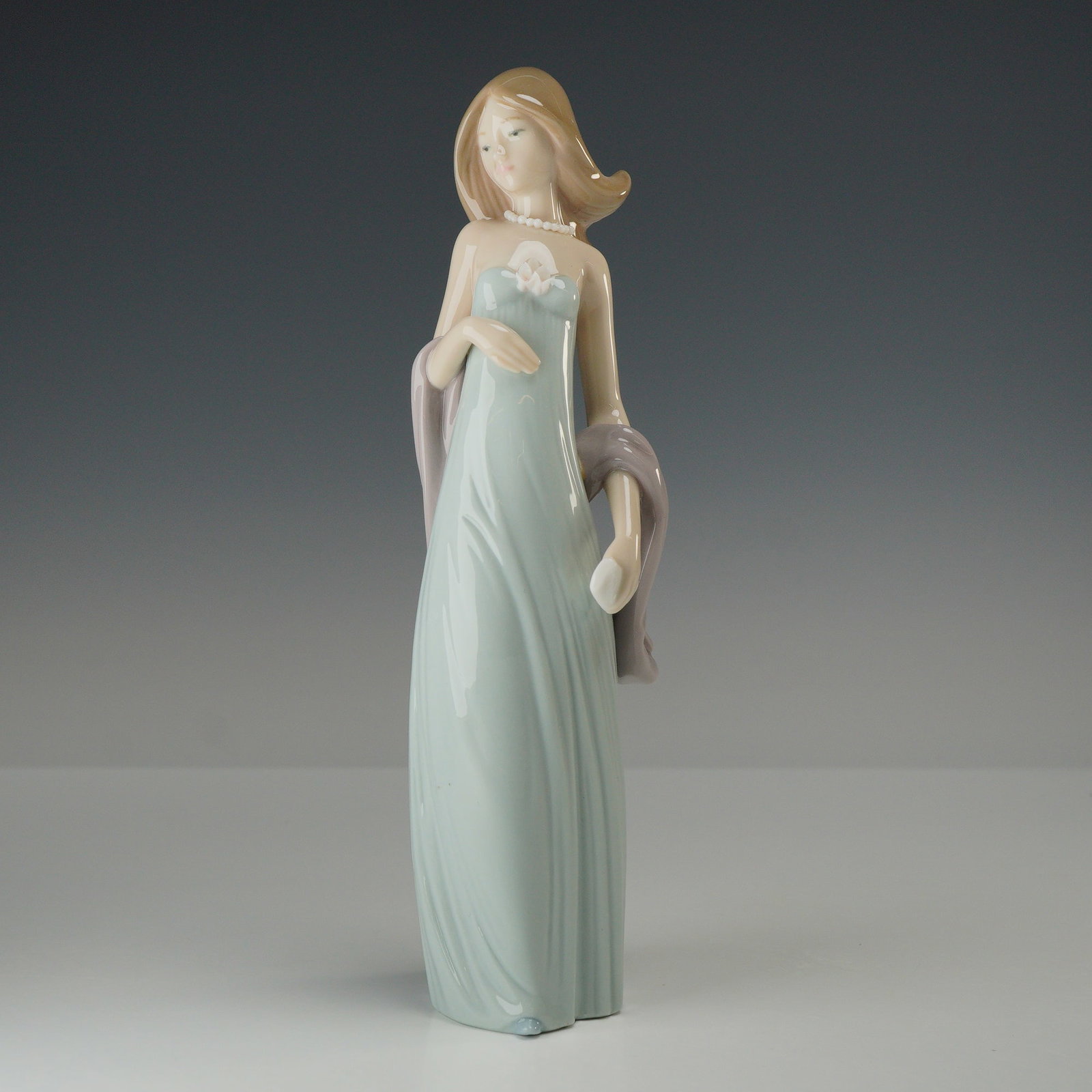 Lladro Ingenue Woman Porcelain Figurine 5487: This piece features a tall, slender female figure in a graceful standing pose. She is depicted wearing a floor-length, sleeveless gown in a pale celadon-blue tone, accented with a delicate white flora