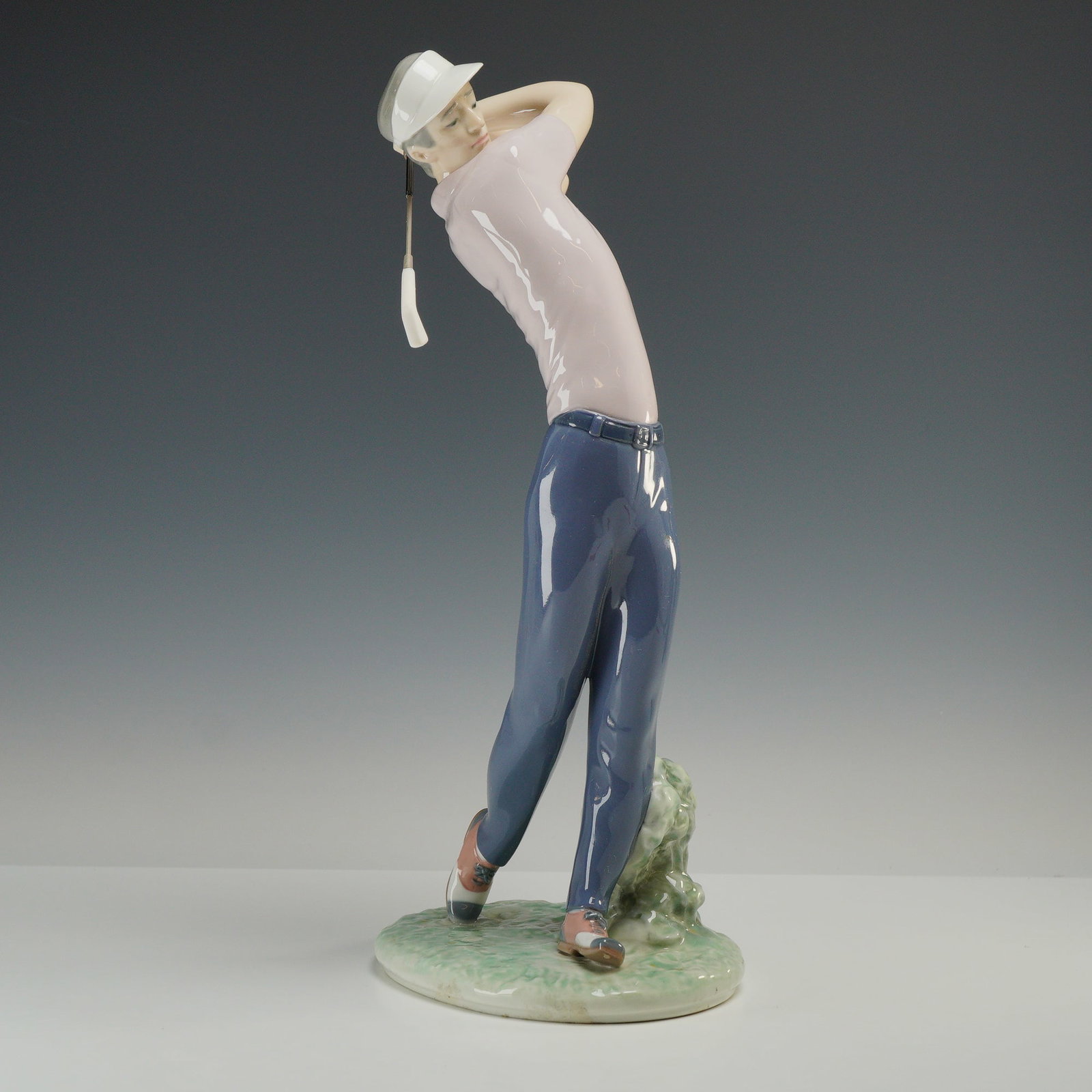 Lladro Male Golfer Porcelain Figurine On the Green 01006032: Lladro On the Green, model number 6032, polychrome glazed porcelain figurine. This piece depicts a male golfer at the completion of his swing, dressed in a light pink short-sleeved polo shirt, dark bl