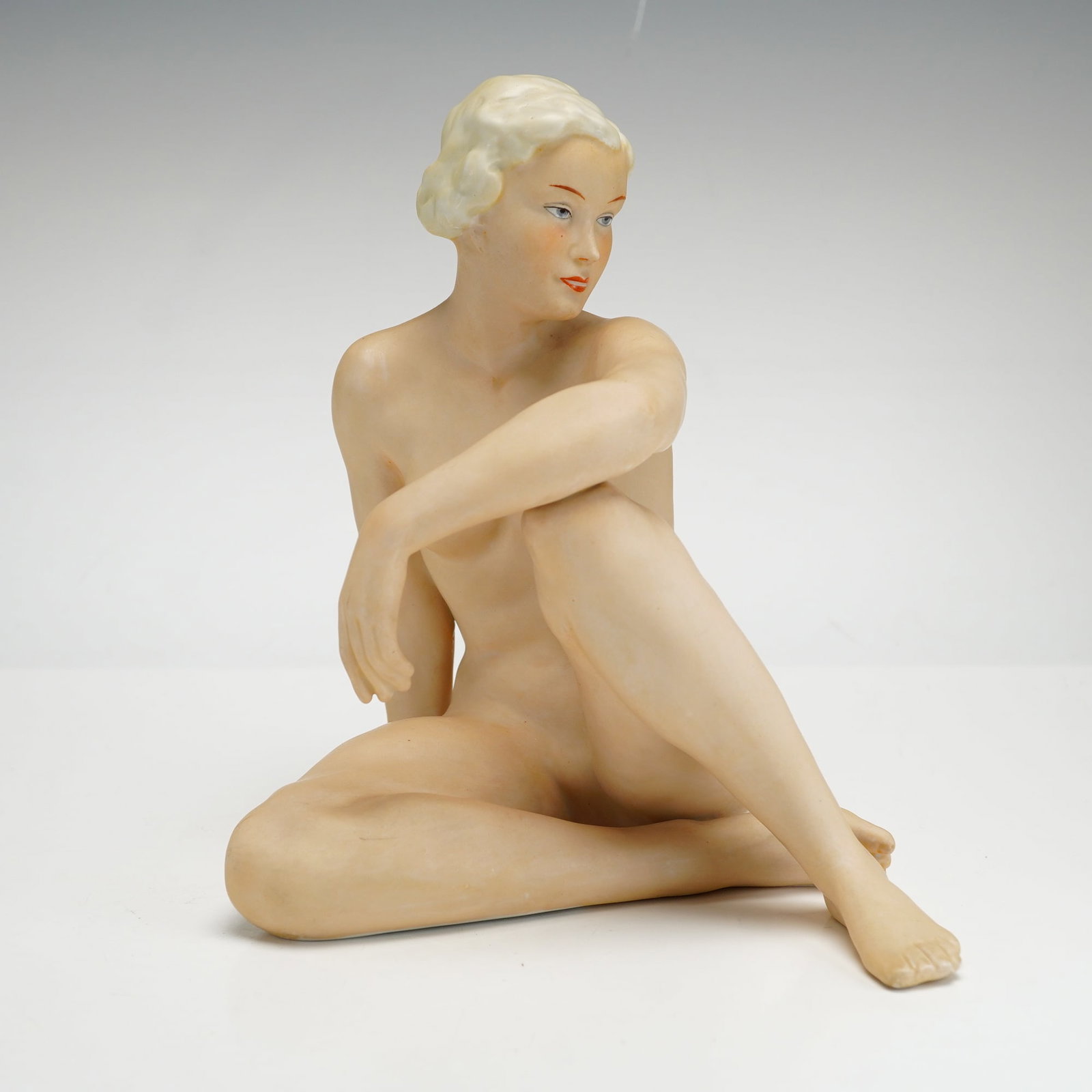 Schaubach Kunst Art Deco Porcelain Figurine, Seated Nude Woman: Schaubach Kunst Art Deco porcelain figurine of a seated nude woman. This piece depicts a female figure with short blonde hair styled in waves, posed with one leg tucked under and the other bent with t