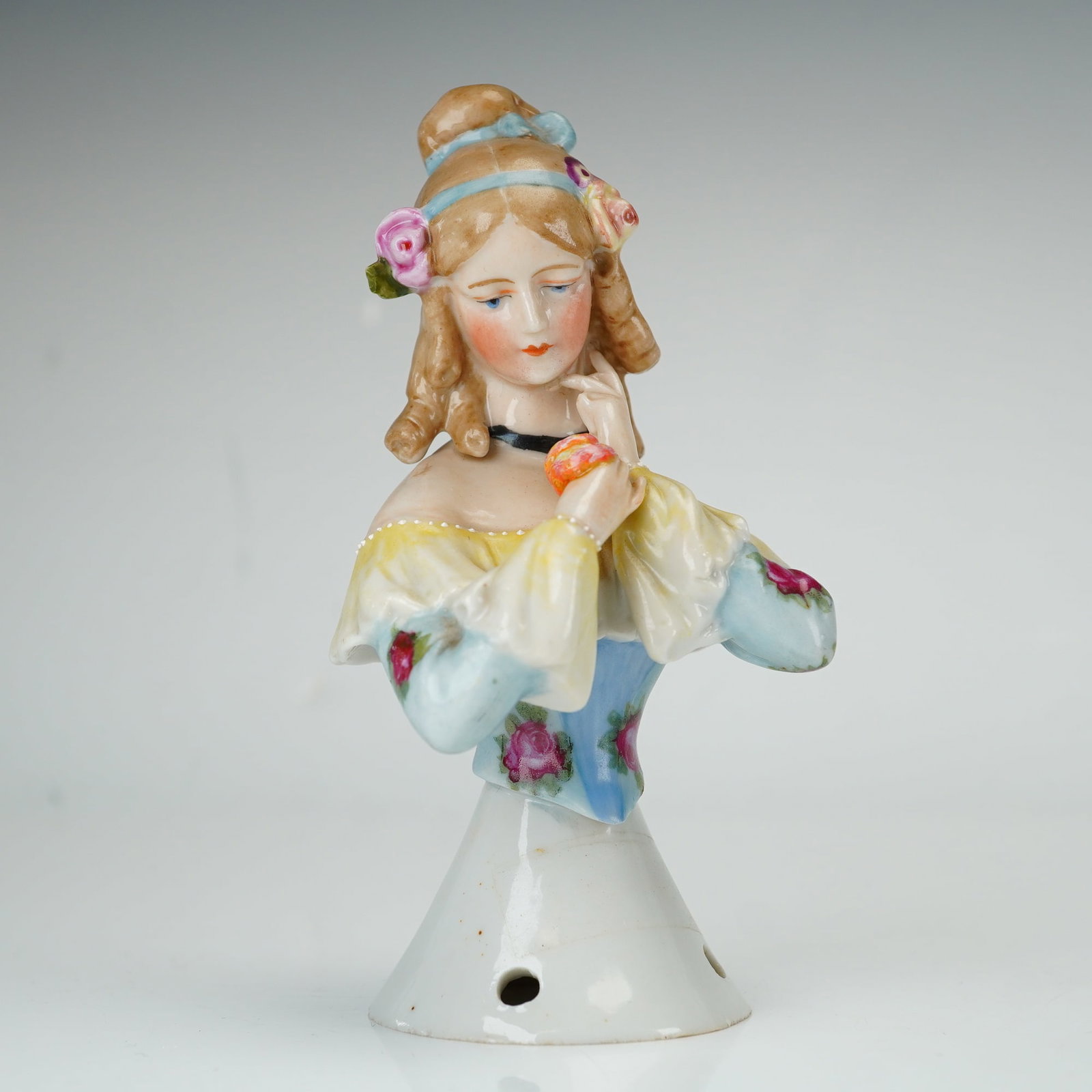 German Porcelain Half Doll Pincushion Figurine: This German polychrome glazed porcelain half doll is designed as a pincushion topper. The figurine depicts a woman from the bust up with light brown hair styled in ringlets and a bun, accented with a 