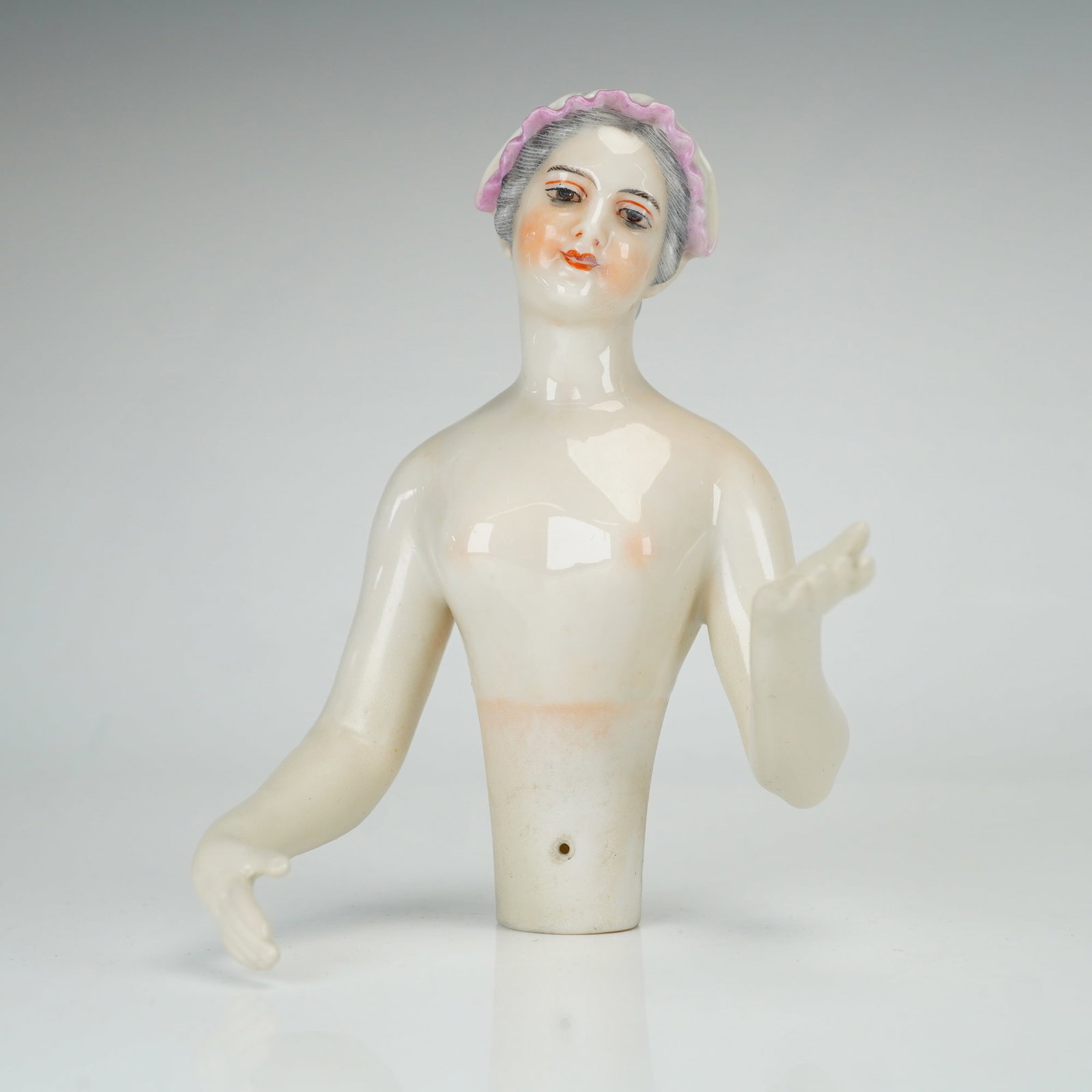 German Porcelain Half Doll by Dressel Kister and Cie: This glazed porcelain half doll was manufactured by Dressel, Kister and Cie in Passau, Germany. The figure depicts a woman with gray hair styled in a bun, wearing a white bonnet with a ruffled pink ri