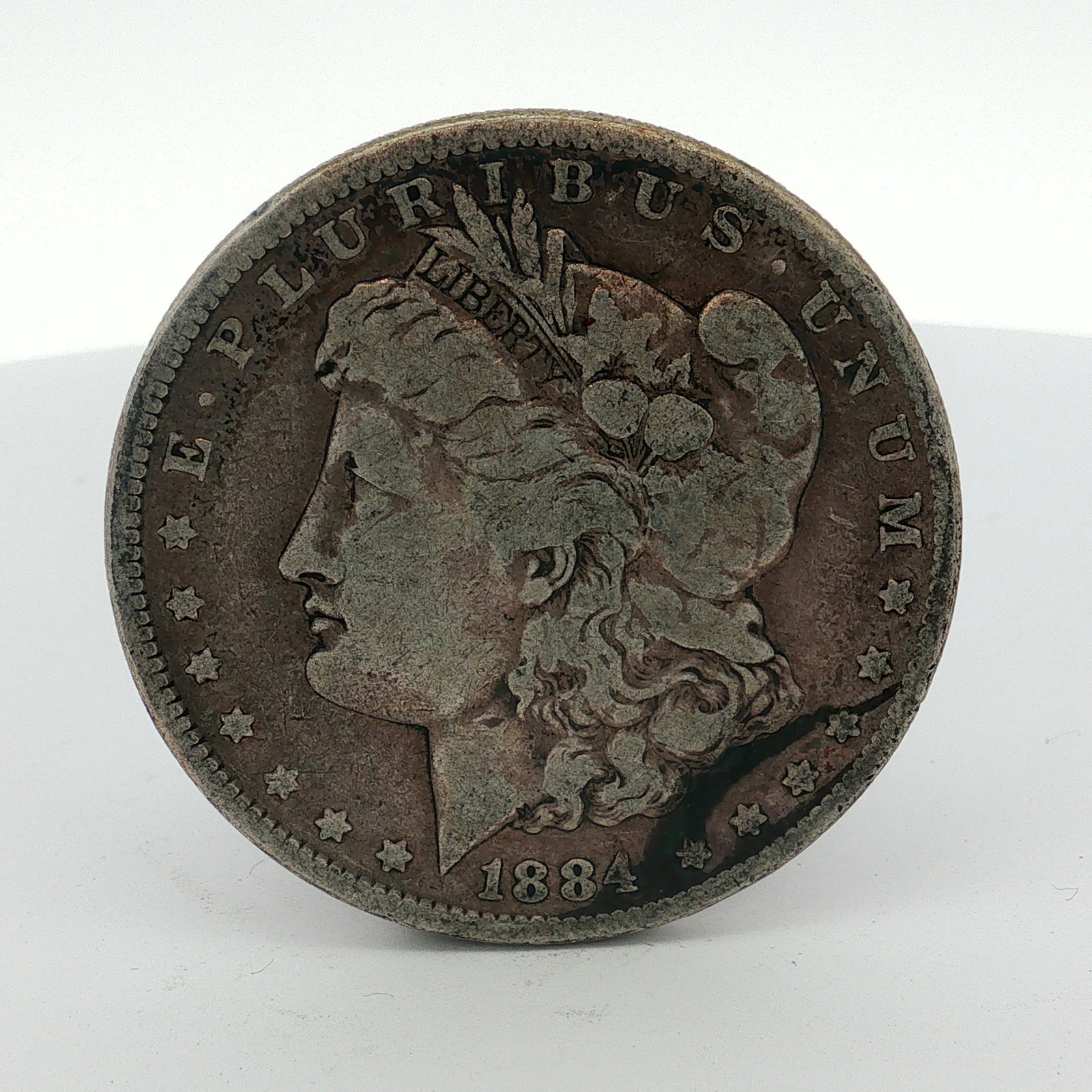1884 US One Dollar Morgan Silver Dollar, Circulated: This Morgan dollar is a US 1884 silver coin struck at the Philadelphia mint. It is composed of 90% silver and 10% copper with a fineness of .900 silver. The obverse features the Liberty Head design ty