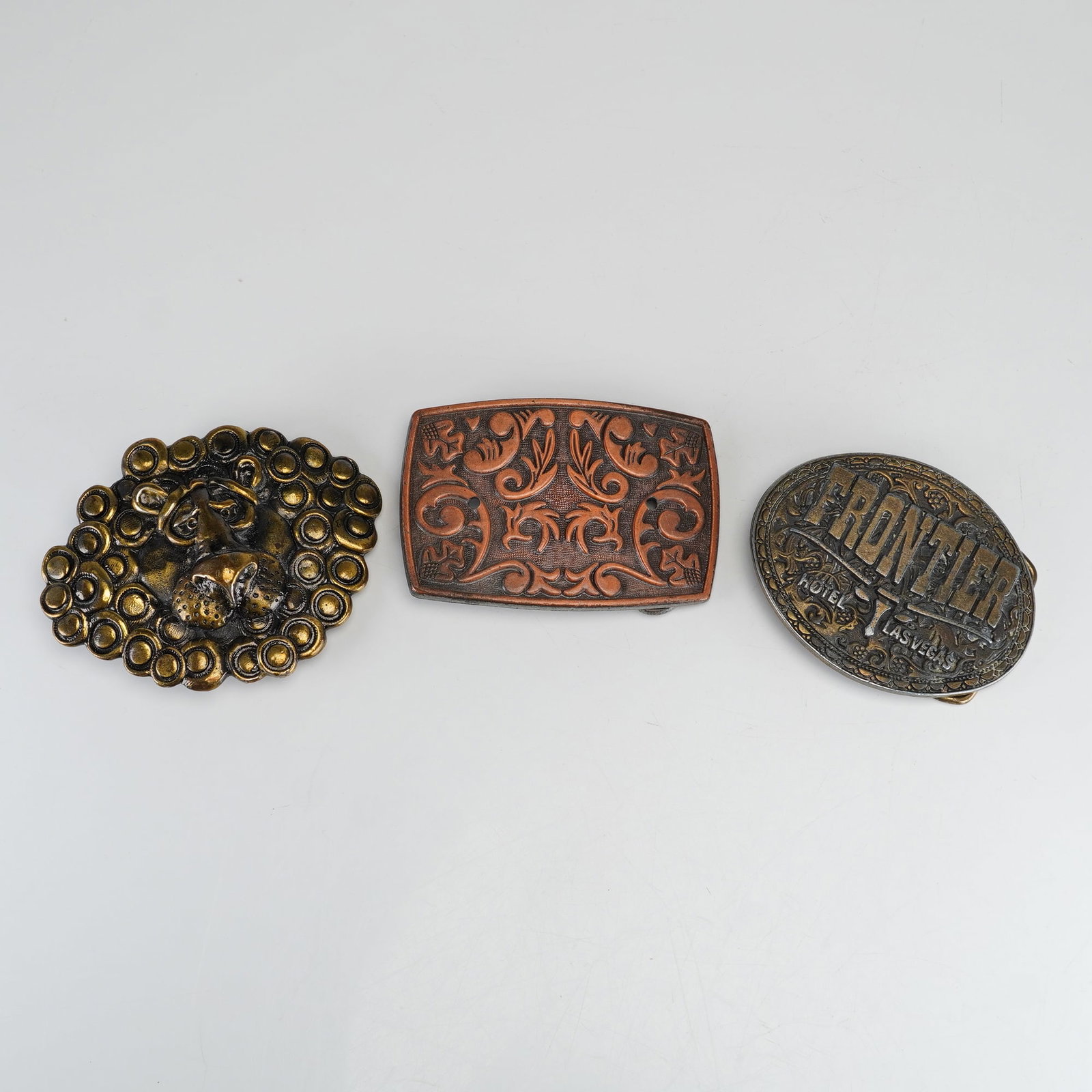 Three Vintage Metal Belt Buckles including The Great American Buckle Co. Lion and Frontier Hotel Las: This lot features three vintage metal belt buckles with various sculptural and embossed designs. The first buckle is a cast metal piece by The Great American Buckle Co. of Chicago, finished in a brass