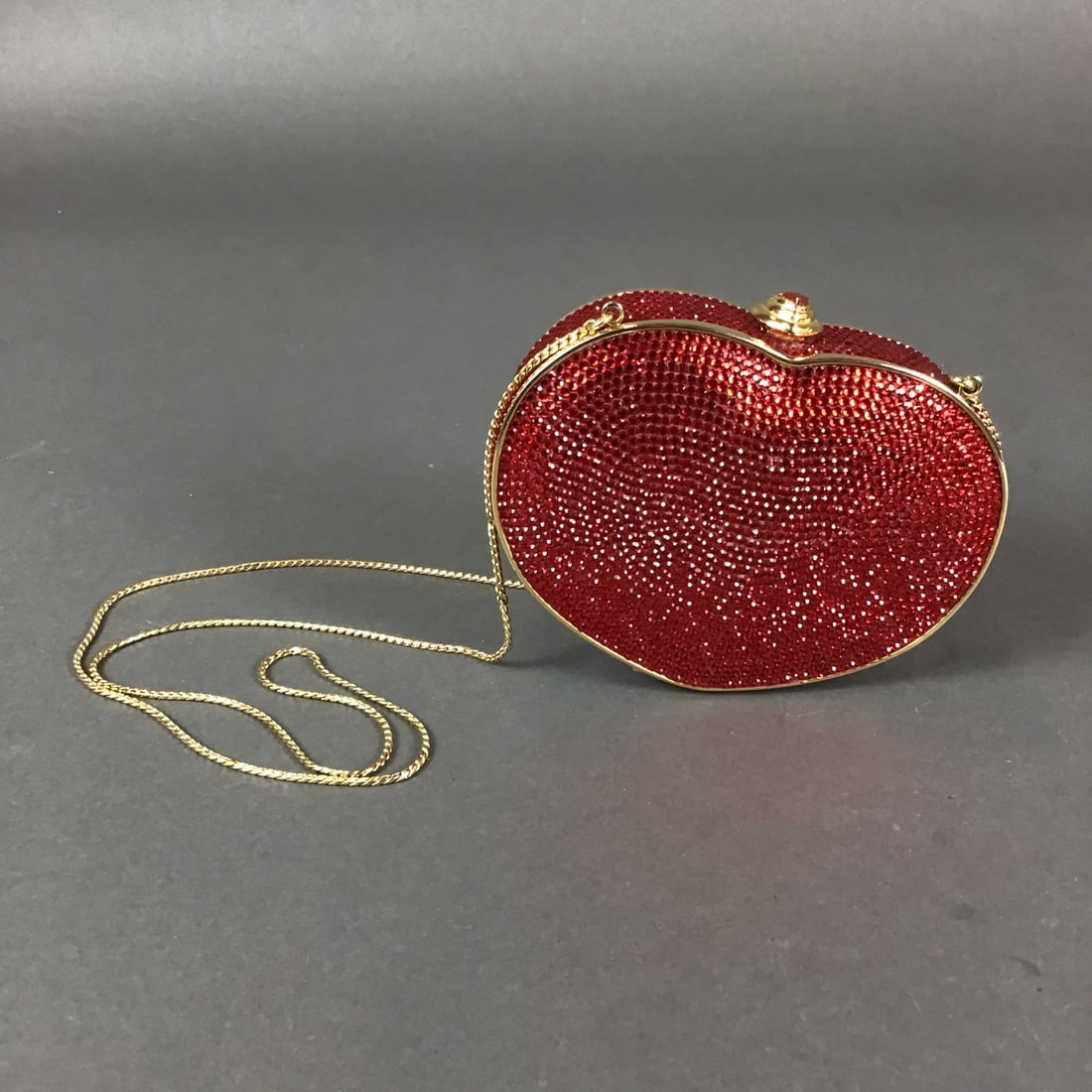 Judith Leiber Rhinestone Red Heart Minaudiere Bag with Gold Chain: This rhinestone heart clutch bag features a hard-shell metal body entirely embellished with pave-set red faceted crystals. The evening bag is designed in a heart-shaped minaudiere style. The exterior