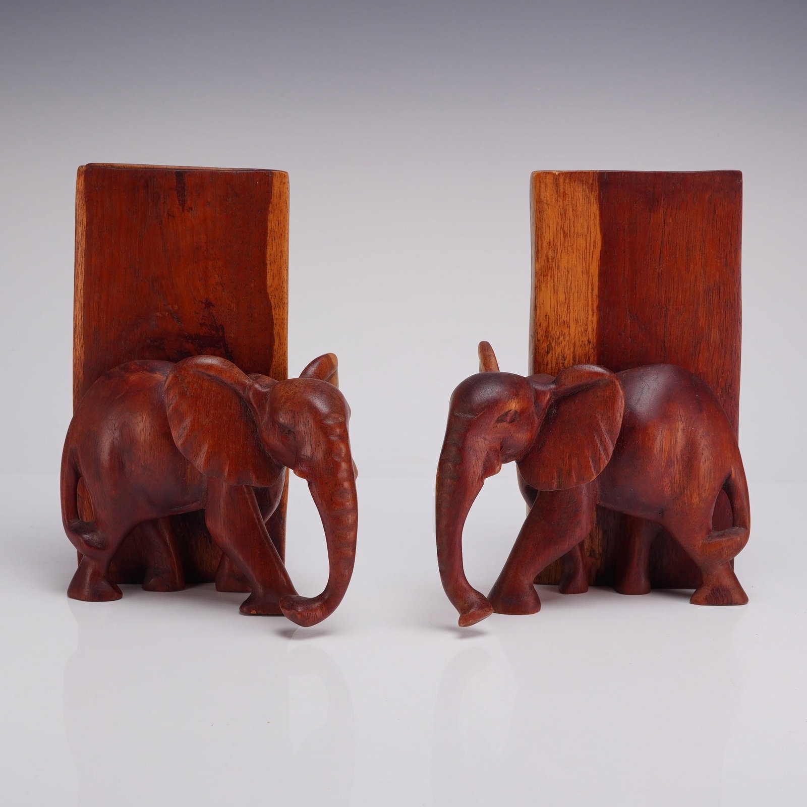 Set of 2 Hand-Carved Wooden Elephant Bookends: Each bookend features a striding elephant carved in relief against a rectangular book-like block, designed in a mirror-image configuration. The items are fashioned from a dense hardwood characterized 