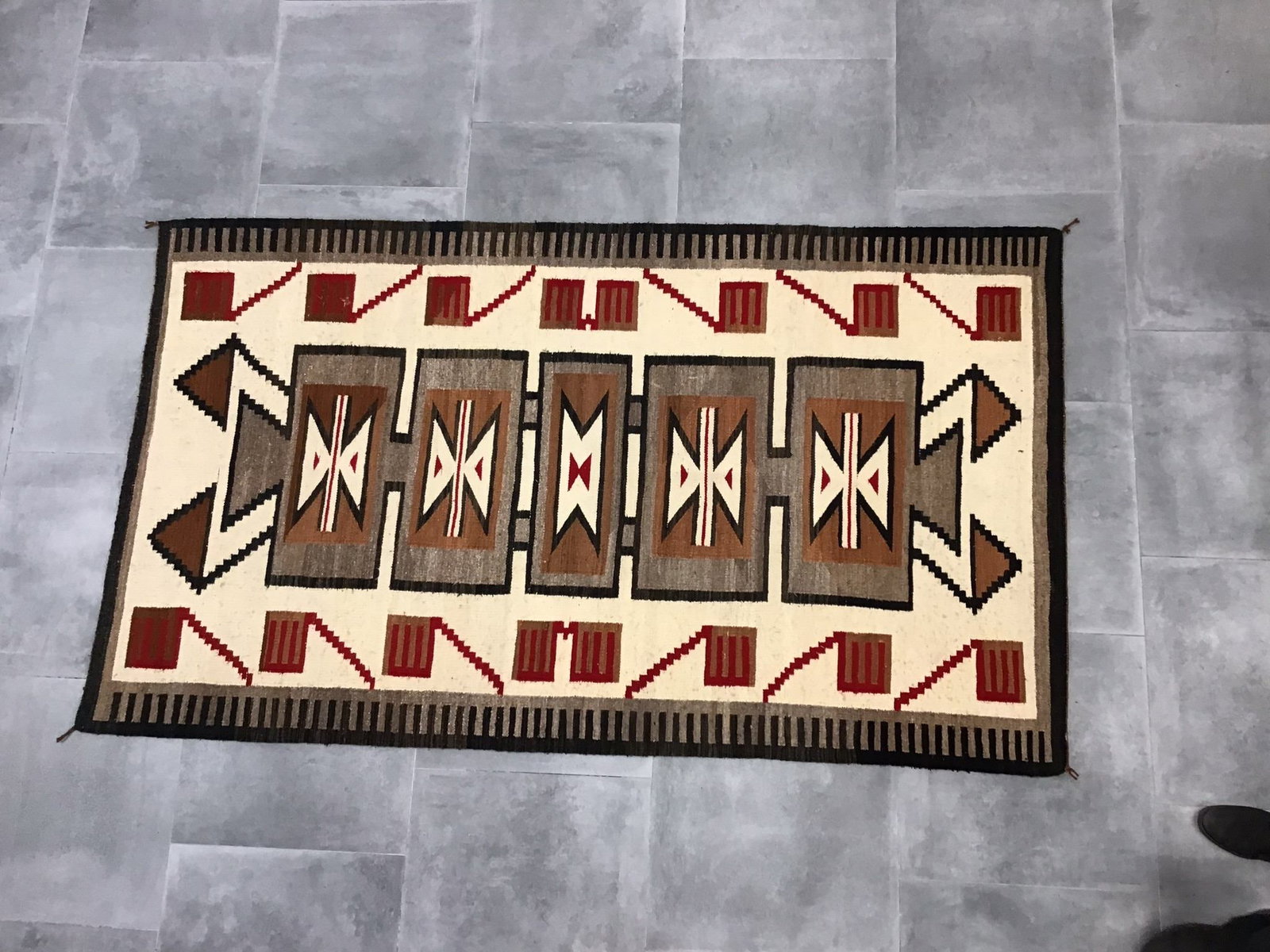Navaho Style Storm Pattern Rug with Red, White, Brown, and Black Accents: A Navaho style storm pattern rug hand-woven in wool. The textile features a central horizontal panel containing five rectangular medallions decorated with geometric bowtie and arrow motifs. The color 
