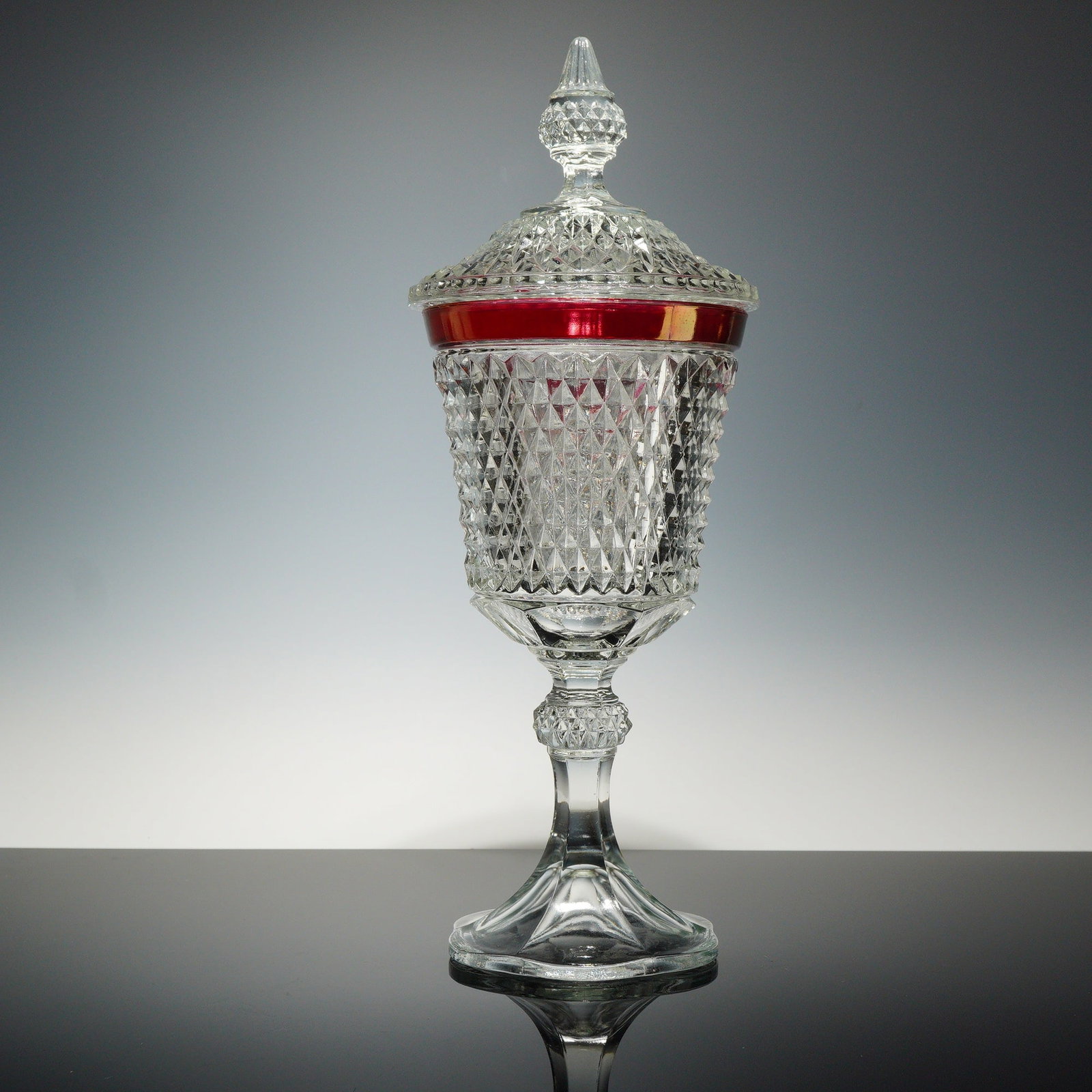 Indiana Glass Diamond Point Ruby Flash Tall Compote Dish with Lid: This tall candy dish is a pressed glass vessel in the Diamond Point pattern by Indiana Glass. The piece consists of a clear glass body and a matching lid, both featuring an overall geometric diamond p