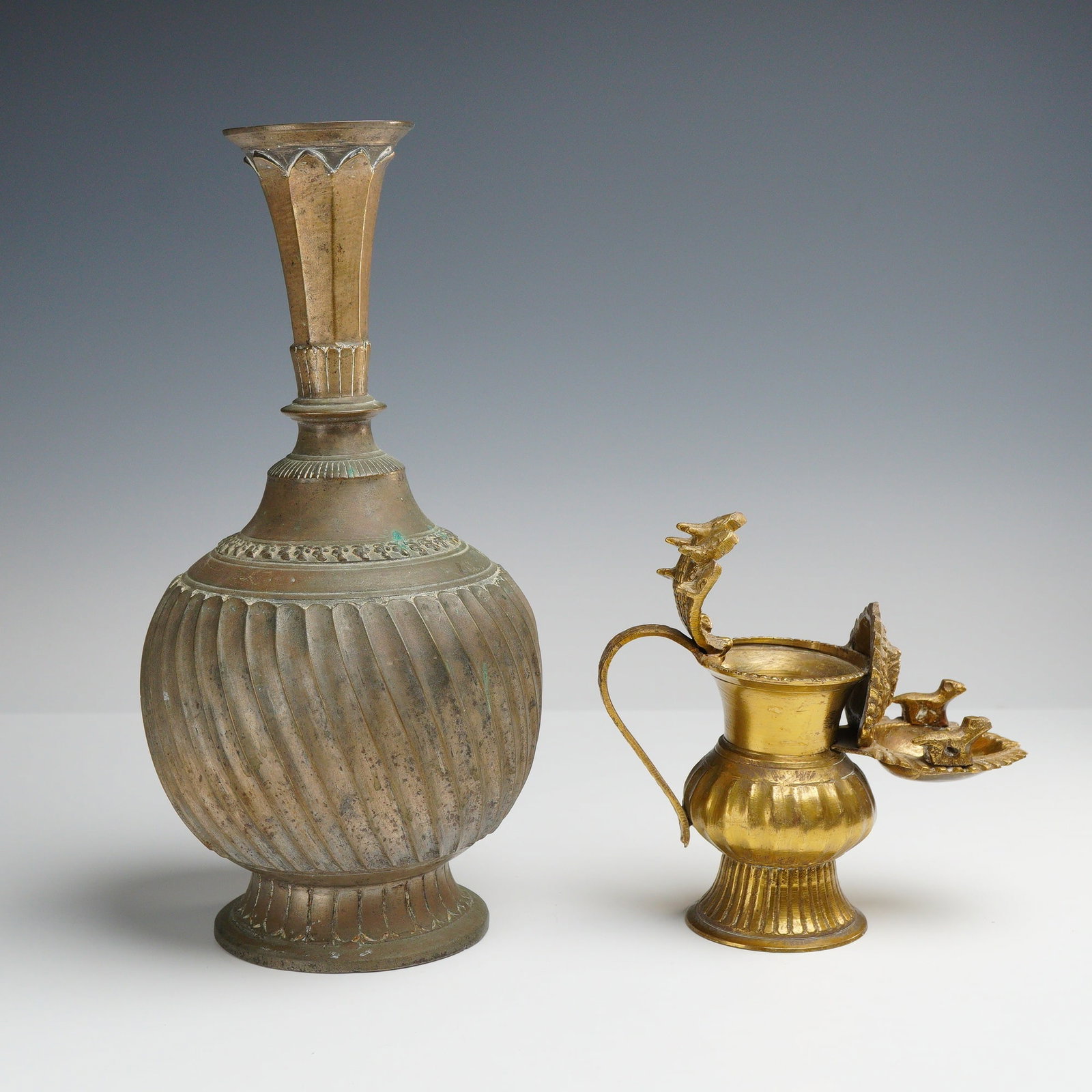 2pc Indian Brass Vessels: This lot comprises two brass vessels of South Asian origin. The first item is a large decorative vessel featuring a globular body with prominent diagonal gadrooning and swirl fluting. The vessel has a