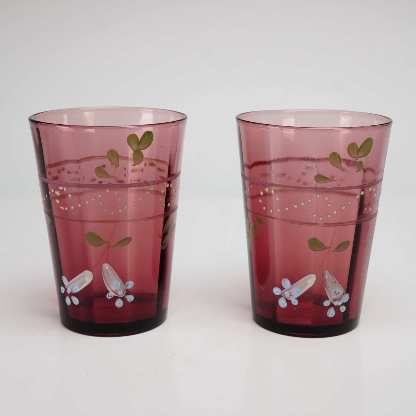 Victorian Bohemian Style Hand-Blown Enameled Cranberry Art Glass Tumblers, Pair: This pair of late 19th to early 20th-century tumblers is likely of Bohemian or French origin. The pieces are hand-blown from transparent cranberry or amethyst-colored glass and feature a subtle molded