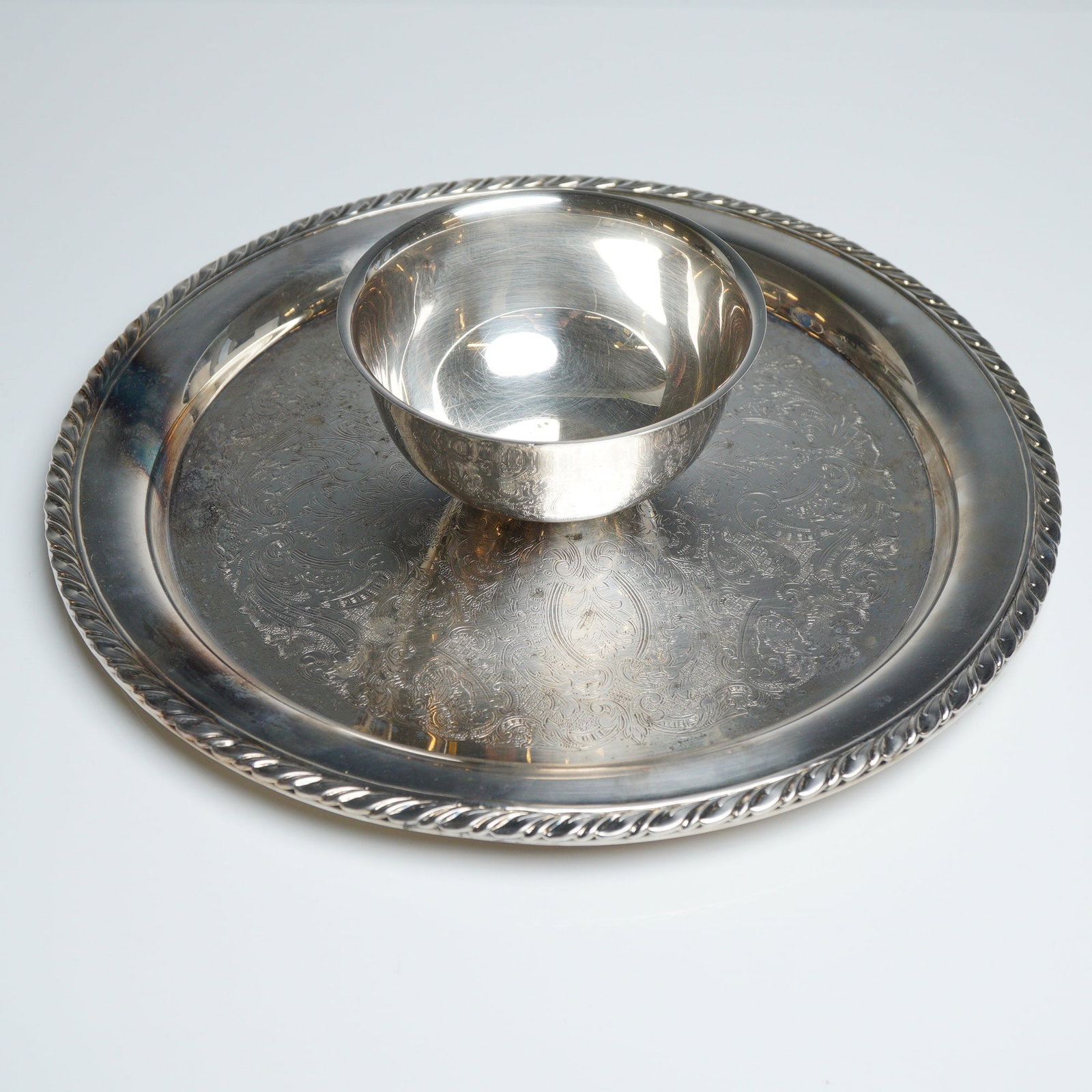 Oneida Silverplated Park Lane Chip and Dip Serving Platter: The piece features a circular tray with an intricately etched floral and foliate center, framed by a raised rim with a decorative rope-twist border. The attached center bowl is of a simple, rounded