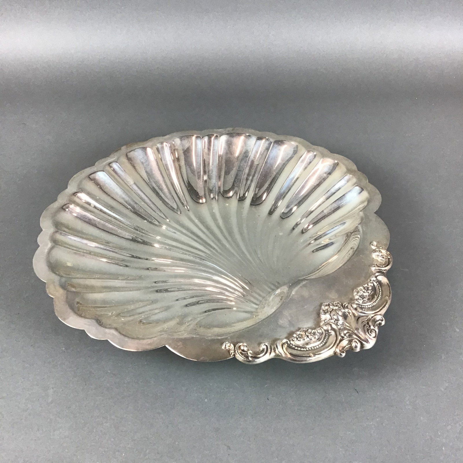 Wallace Baroque Silverplate Shell Form Dish: A silverplate dish in the Baroque pattern by Wallace Silversmiths. The object features a fluted and scalloped shell-shaped body with an ornate handle decorated with rococo-style scrolls and floral
