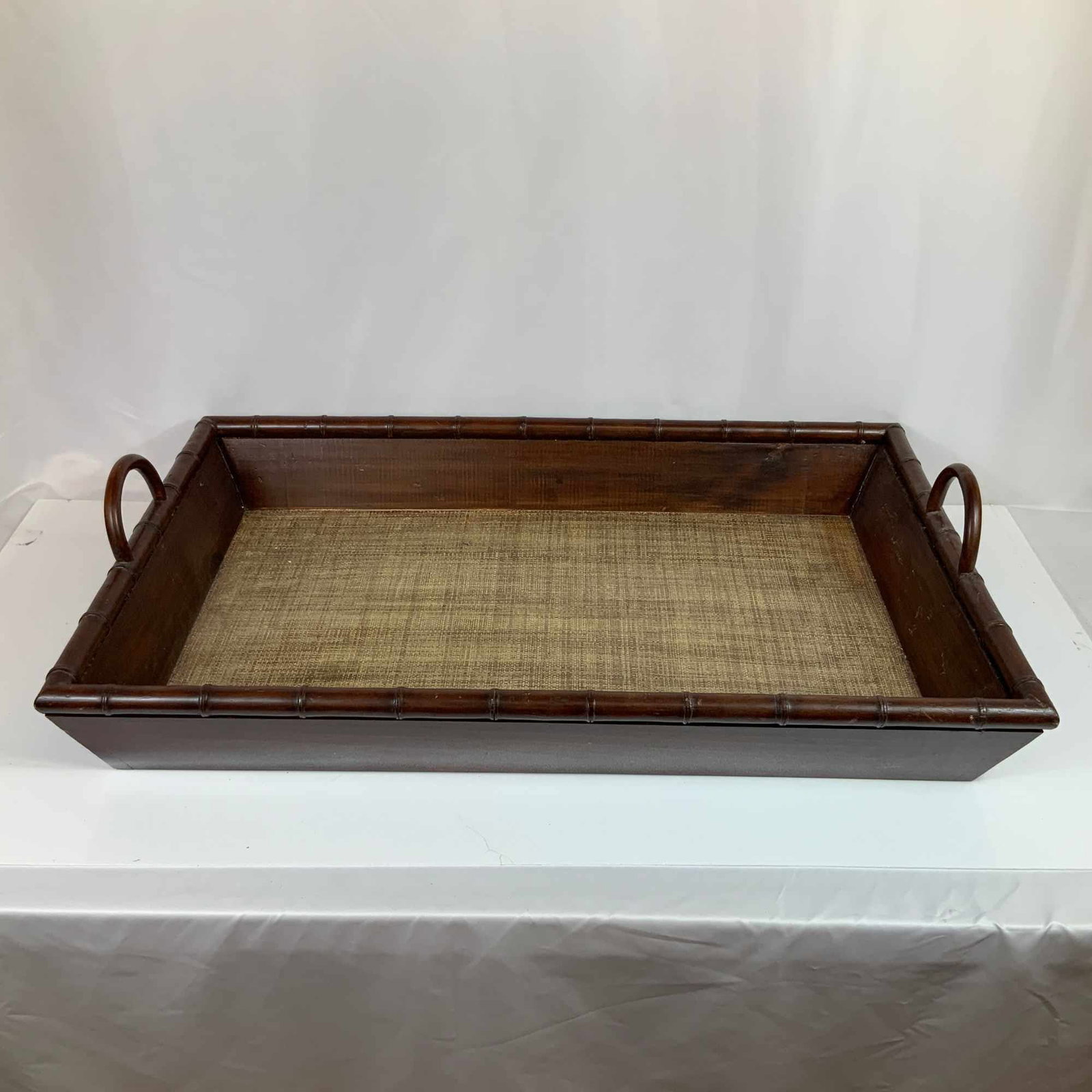 British Colonial Style Faux Bamboo and Grasscloth Wooden Serving Tray: A rectangular wooden serving tray featuring a faux bamboo carved rim and integrated loop handles. The tray is finished in a dark brown stain, with the interior base lined with a woven grasscloth or