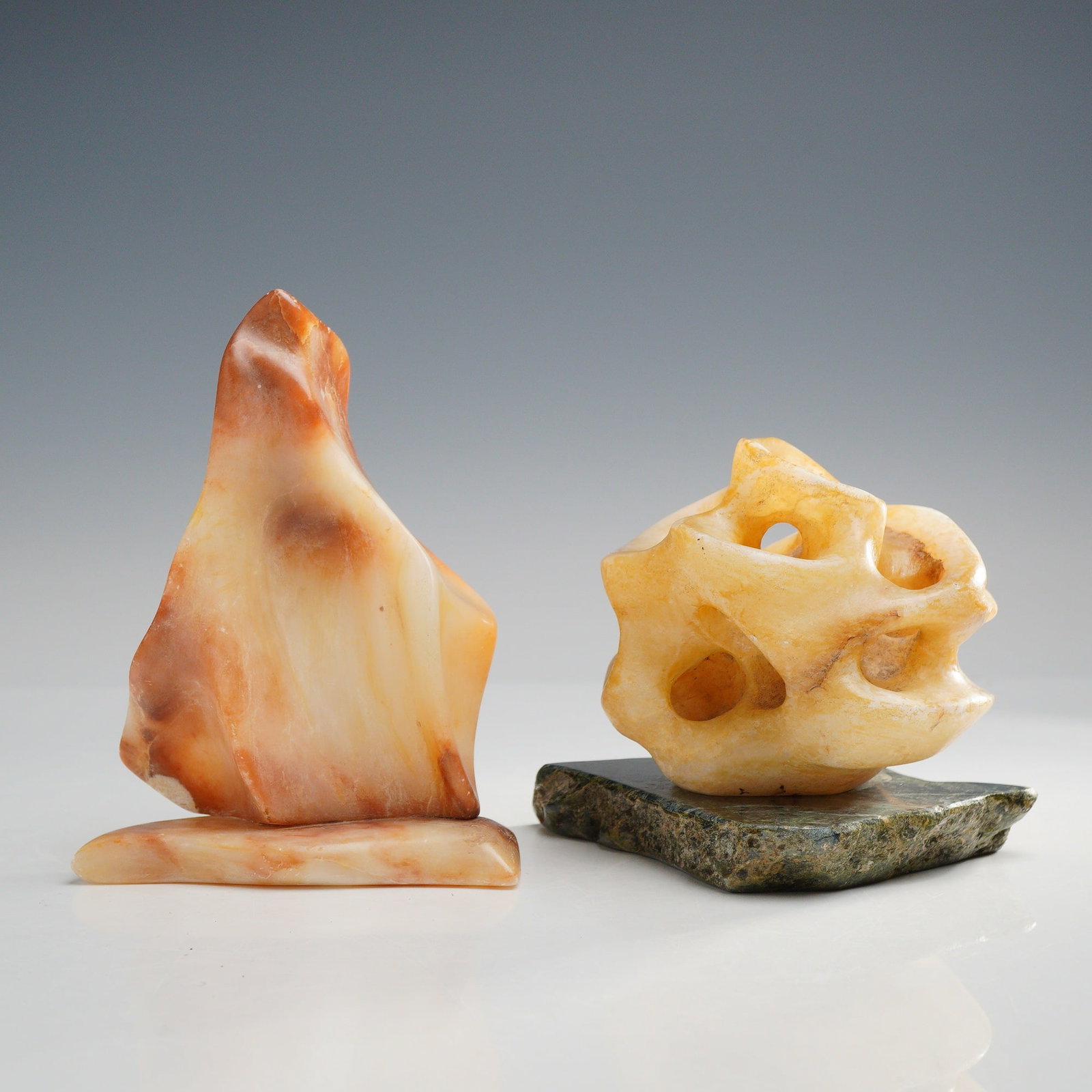 2pc Mid-Century Modern Abstract Carved Stone Rock Sculptures: A pair of mid-century modern abstract stone sculptures of biomorphic form. The first piece is carved from a marbled reddish-orange and cream-colored stone, with characteristics consistent with carneli