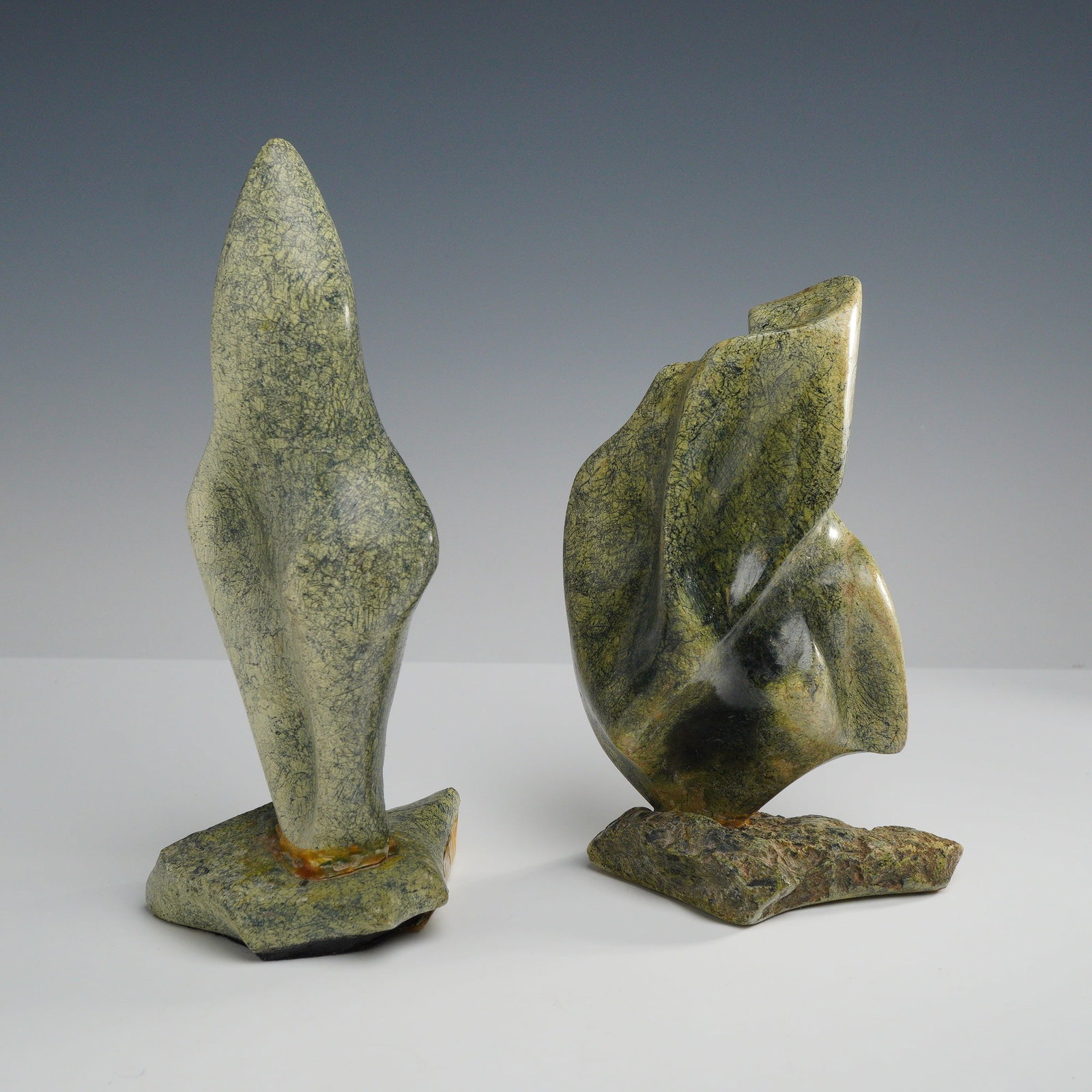 2pc Modernist Abstract Carved Leopard Rock Sculptures: A pair of Modernist abstract biomorphic sculptures carved from mottled greenish leopard rock. The first sculpture features a tall, slender, and tapering form with a smooth, polished surface. The secon