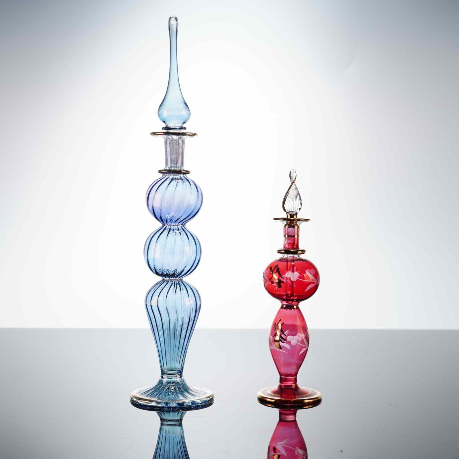 2pc Decorative Art Glass Bottles, Blue and Cranberry Egyptian Style: This lot features a pair of decorative art glass bottles. The larger bottle is crafted from transparent blue glass with a light blue gradient, featuring optic-molded ribbing across three bulbous secti