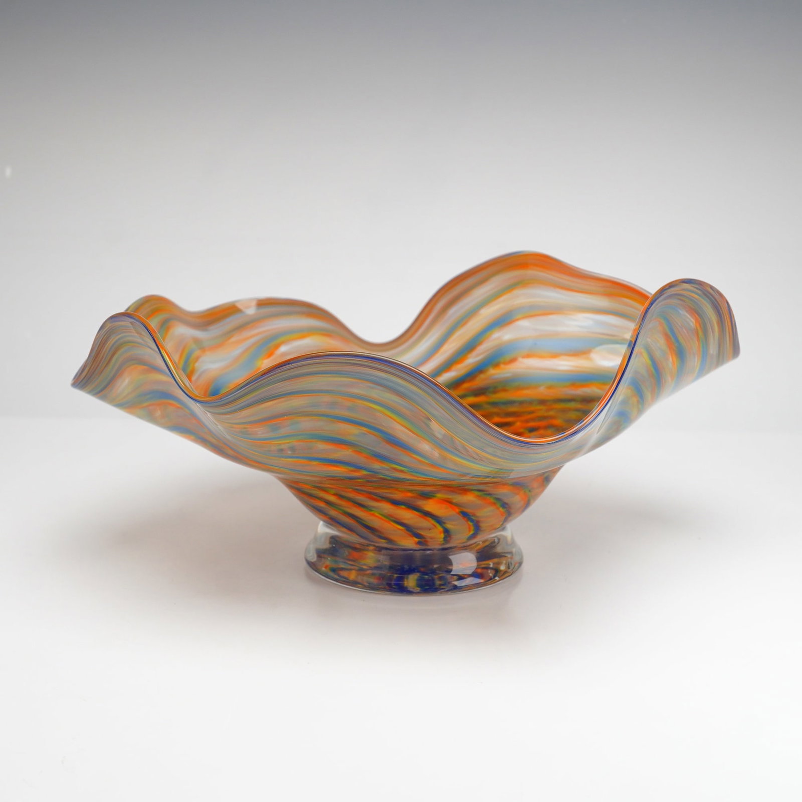 Multi-Colored Studio Art Glass Centerpiece Wave Bowl: This hand-blown studio art glass bowl features an undulating wavy rim with a spiraling pattern of multi-colored glass canes in shades of orange, cobalt blue, yellow, and red. The decoration consists o